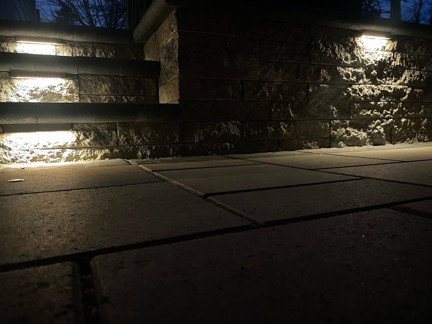 Stone steps and walkway illuminated by soft lights at night, creating a warm and inviting outdoor ambiance. Trees are visible in the background.