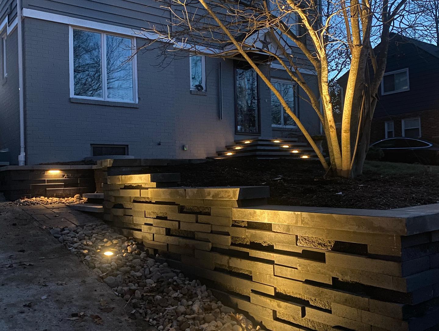 Modern house entrance at dusk, featuring illuminated stone pathway and decorative lighting. A leafless tree adds an artistic touch to the landscape.