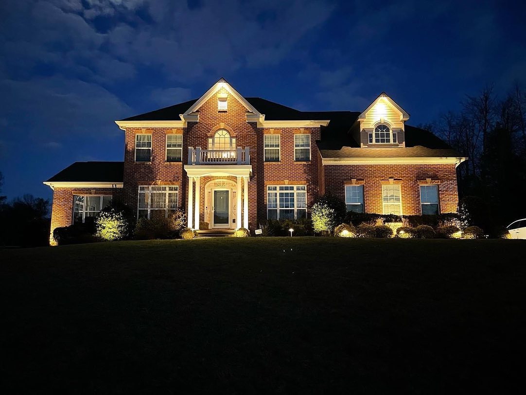 Brick house illuminated at night, featuring multiple windows, a central door, and landscaped bushes in front, under a dark, cloudy sky.