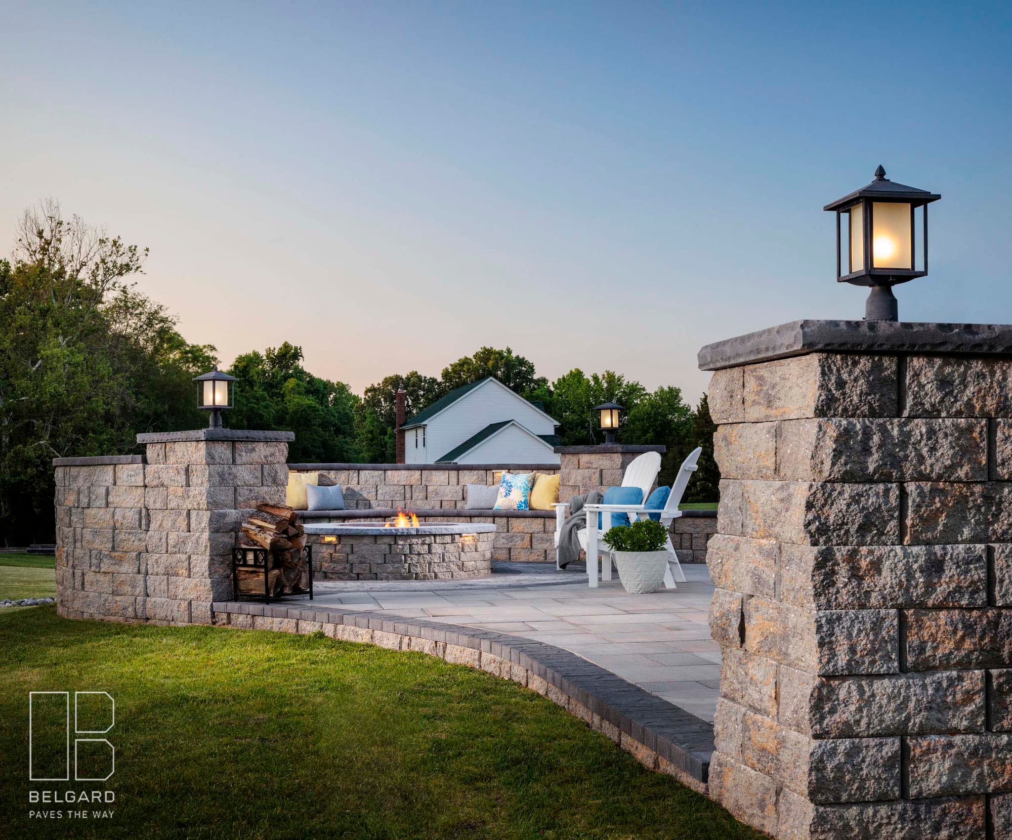 A cozy outdoor patio with stone seating, a fire pit, and elegant lanterns. Surrounded by grass and trees, under a clear sky.