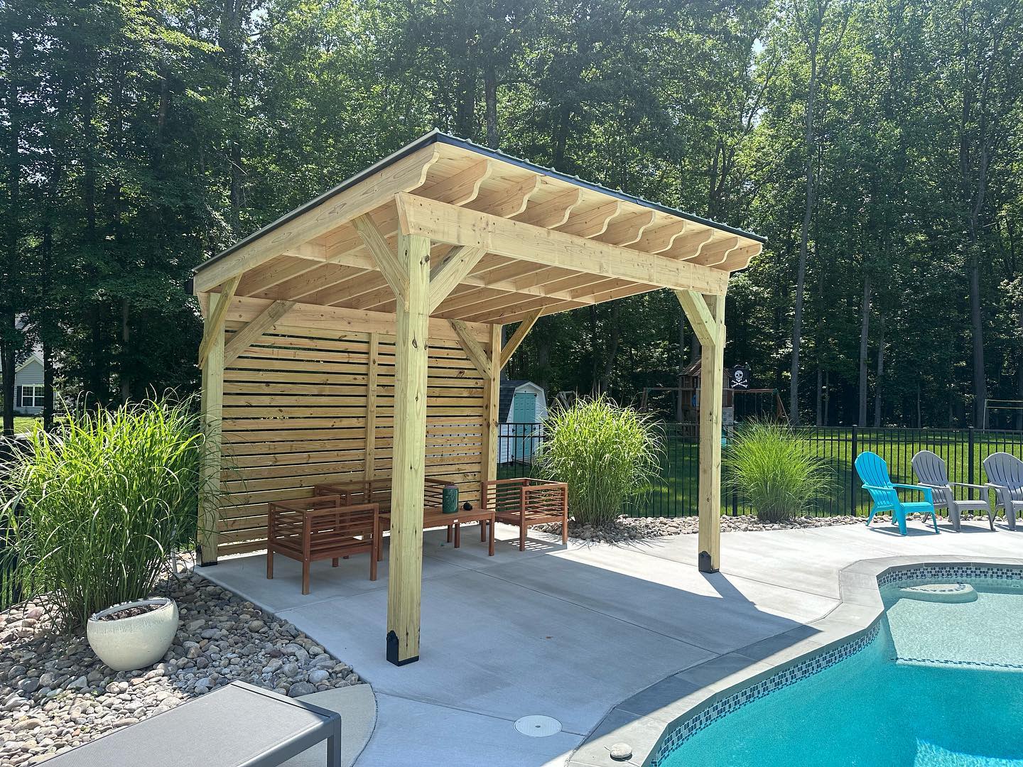 A wooden gazebo overlooks a swimming pool surrounded by lush greenery and colorful chairs, creating a serene backyard retreat.