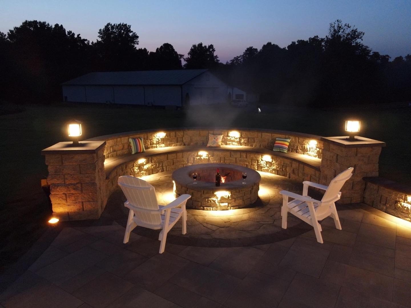 An outdoor patio with fire pit, surrounded by illuminated stone seating, two empty Adirondack chairs, and trees in the background at dusk.
