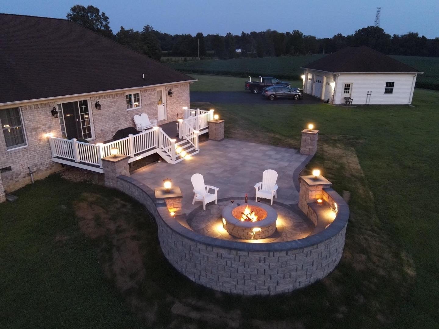 A cozy patio with a fire pit, surrounded by illuminated stone wall and chairs. Adjacent house and garage in a spacious yard.