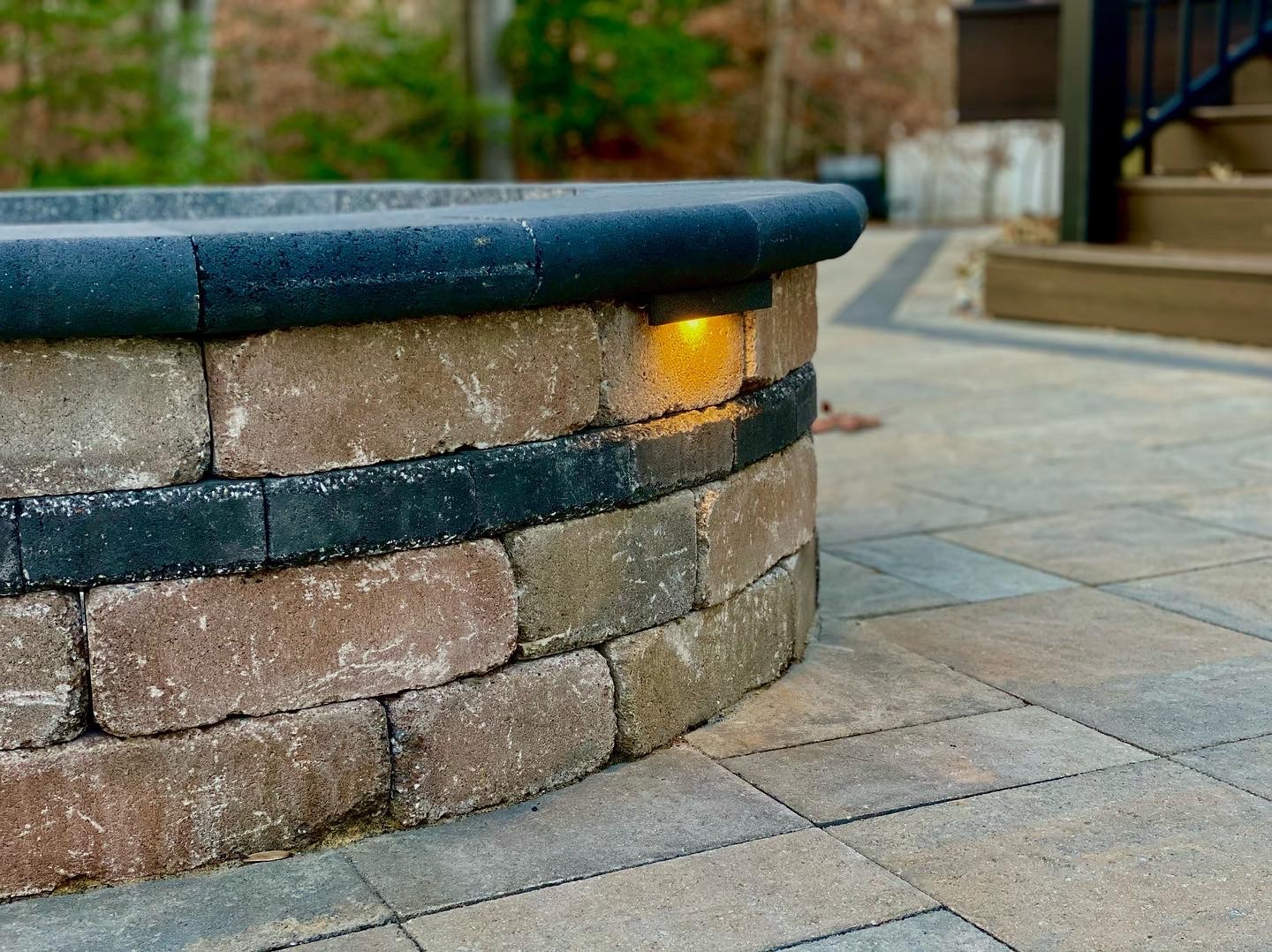 Circular stone fire pit with a small embedded light, surrounded by paved patio. Background includes steps and blurred trees.
