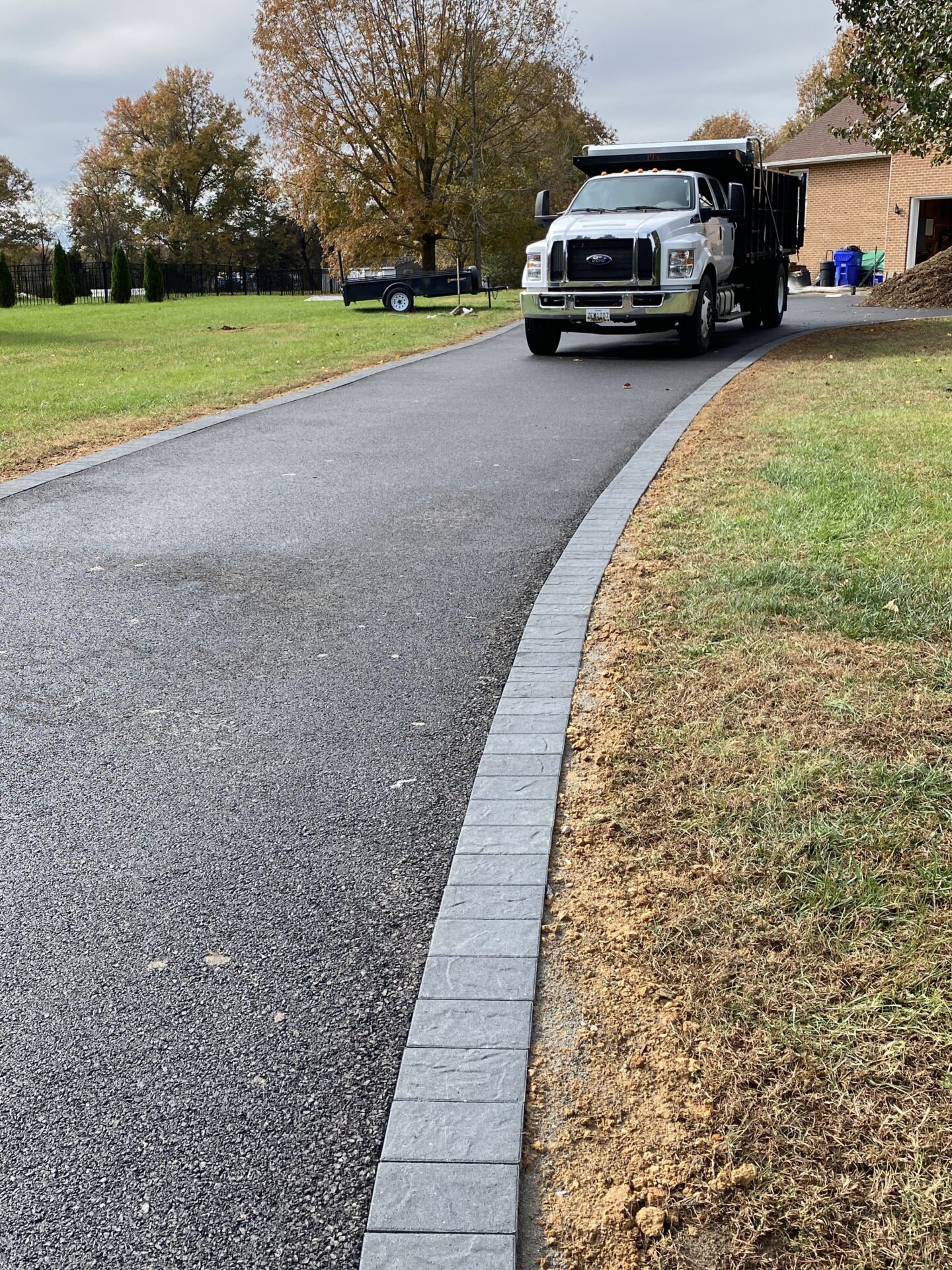 A dump truck is parked on a newly paved driveway beside a brick building; trees and a small trailer are in the background.