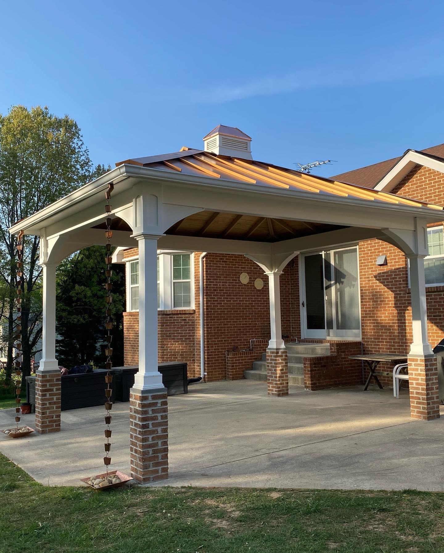 A backyard with a brick house features a covered patio, wooden table, chairs, and a rain chain, surrounded by grassy lawn and trees.