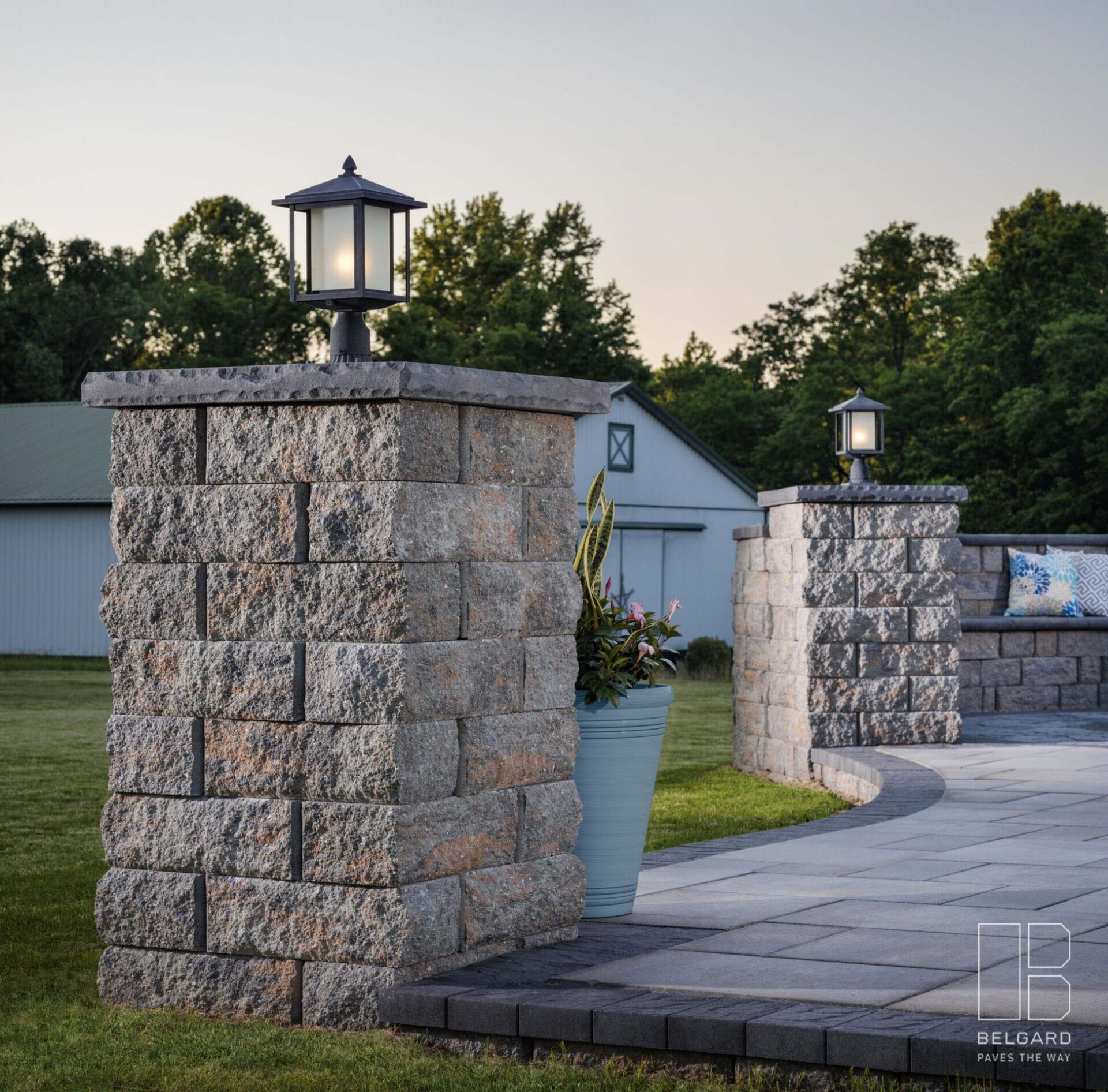 Stone pillars with lanterns line a curved pathway. A light blue planter with flowers sits nearby, and a barn is visible in the background.