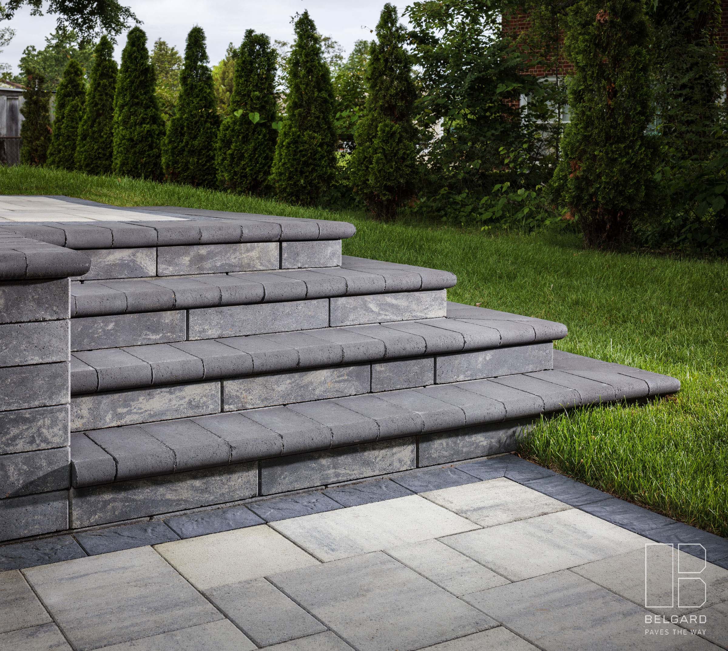 Stone steps lead to a grassy area surrounded by tall trees, featuring Belgard branding in the corner of the image.