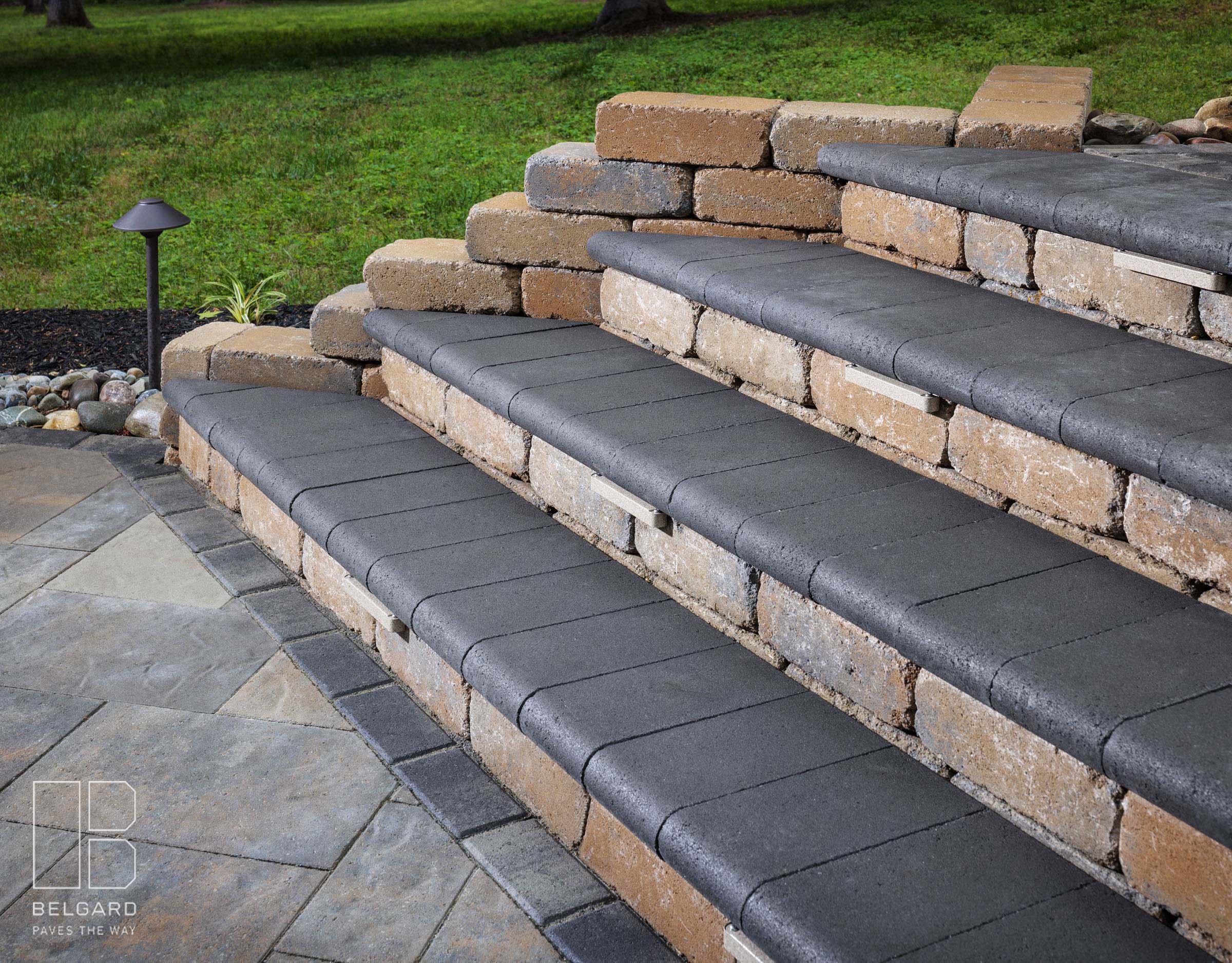 Stone steps with dark caps lead to a grassy area. Decorative rock garden and single outdoor light fixture on the side.