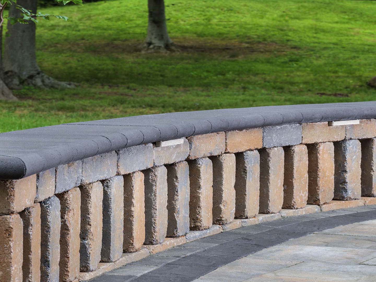 Curved stone wall with a smooth top, set against a grassy landscape with trees in the background at a park setting.