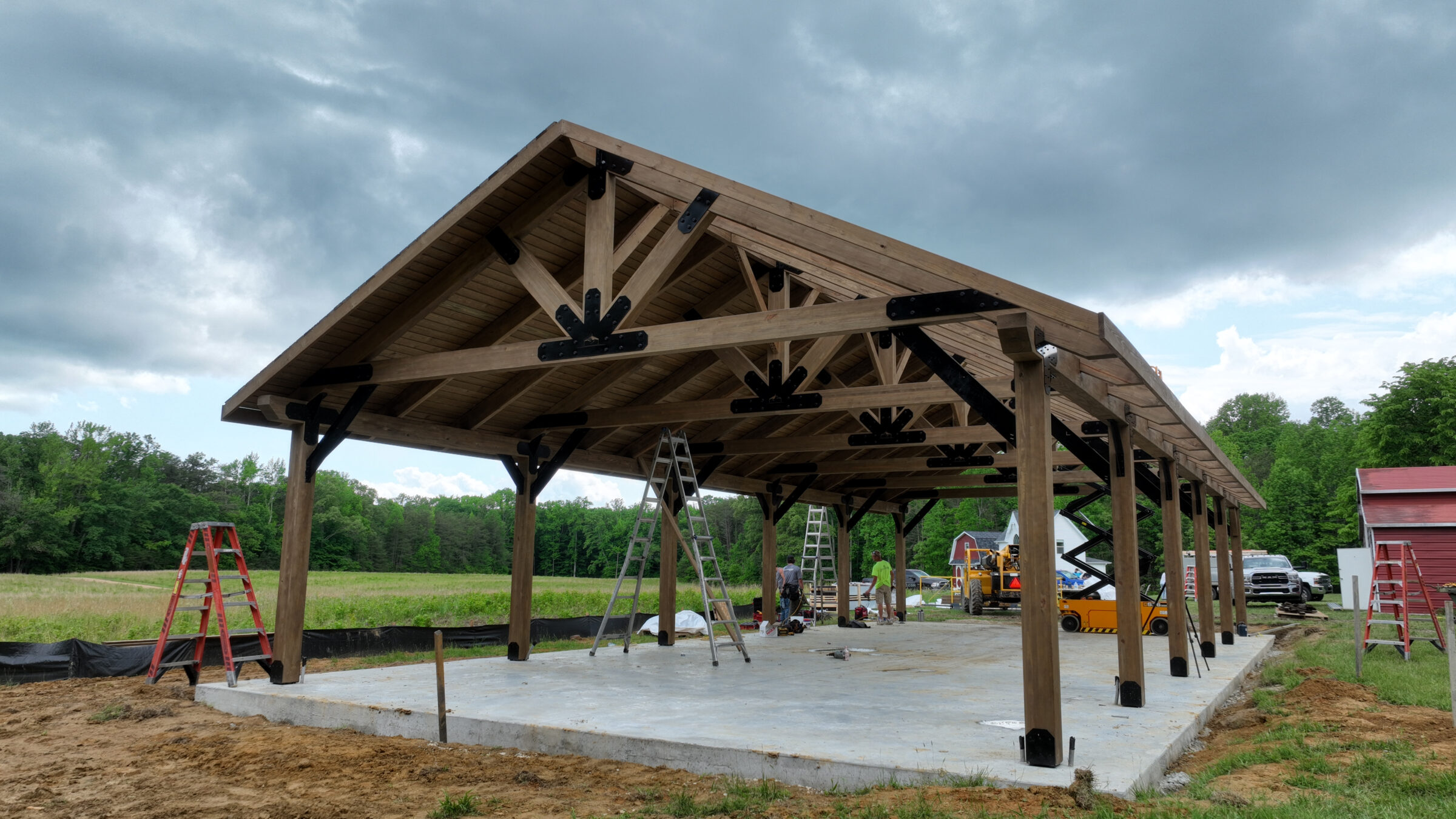 Wooden pavilion under construction on concrete slab, surrounded by ladders and equipment, with a forest backdrop under a cloudy sky.