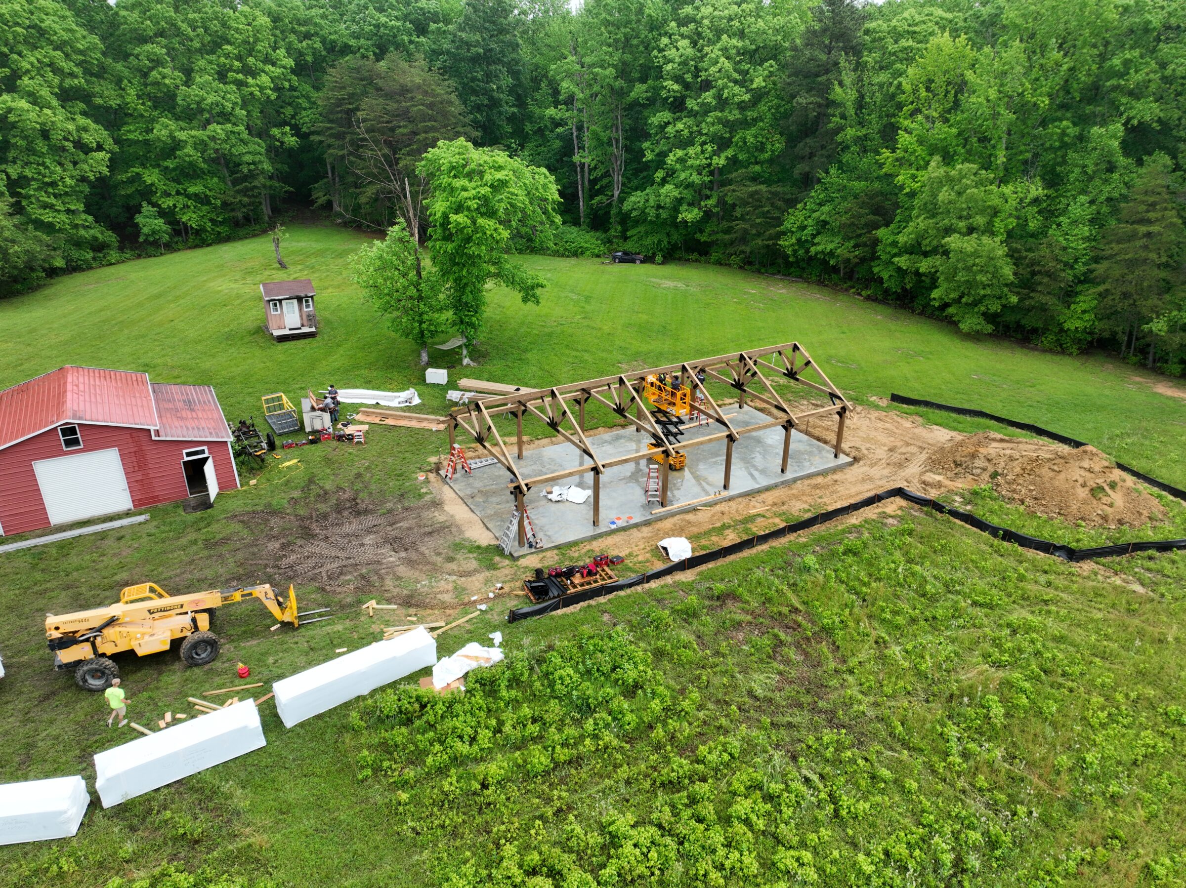 A construction site in a grassy area with a partially built structure, red shed, machinery, and people working alongside wooded surroundings.