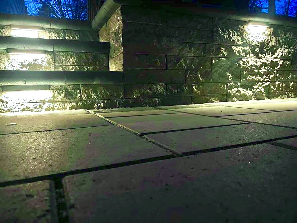 Stone steps and patio lit by embedded lights, surrounded by low walls. Trees are visible in the background against an evening sky.