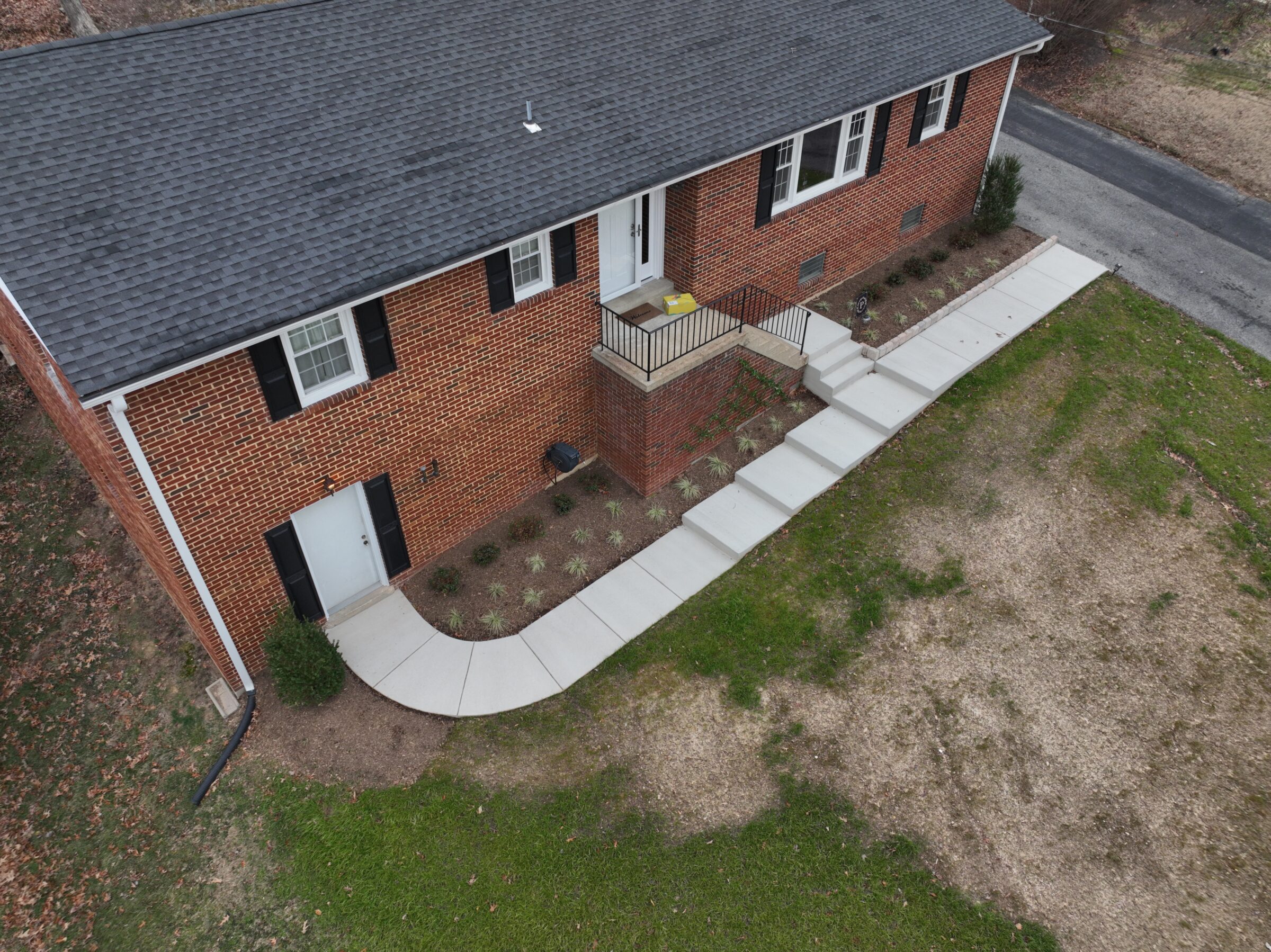 Aerial view of a large brick house with black shutters, white doors, neat landscaping, and a concrete walkway. No visible people or landmarks.