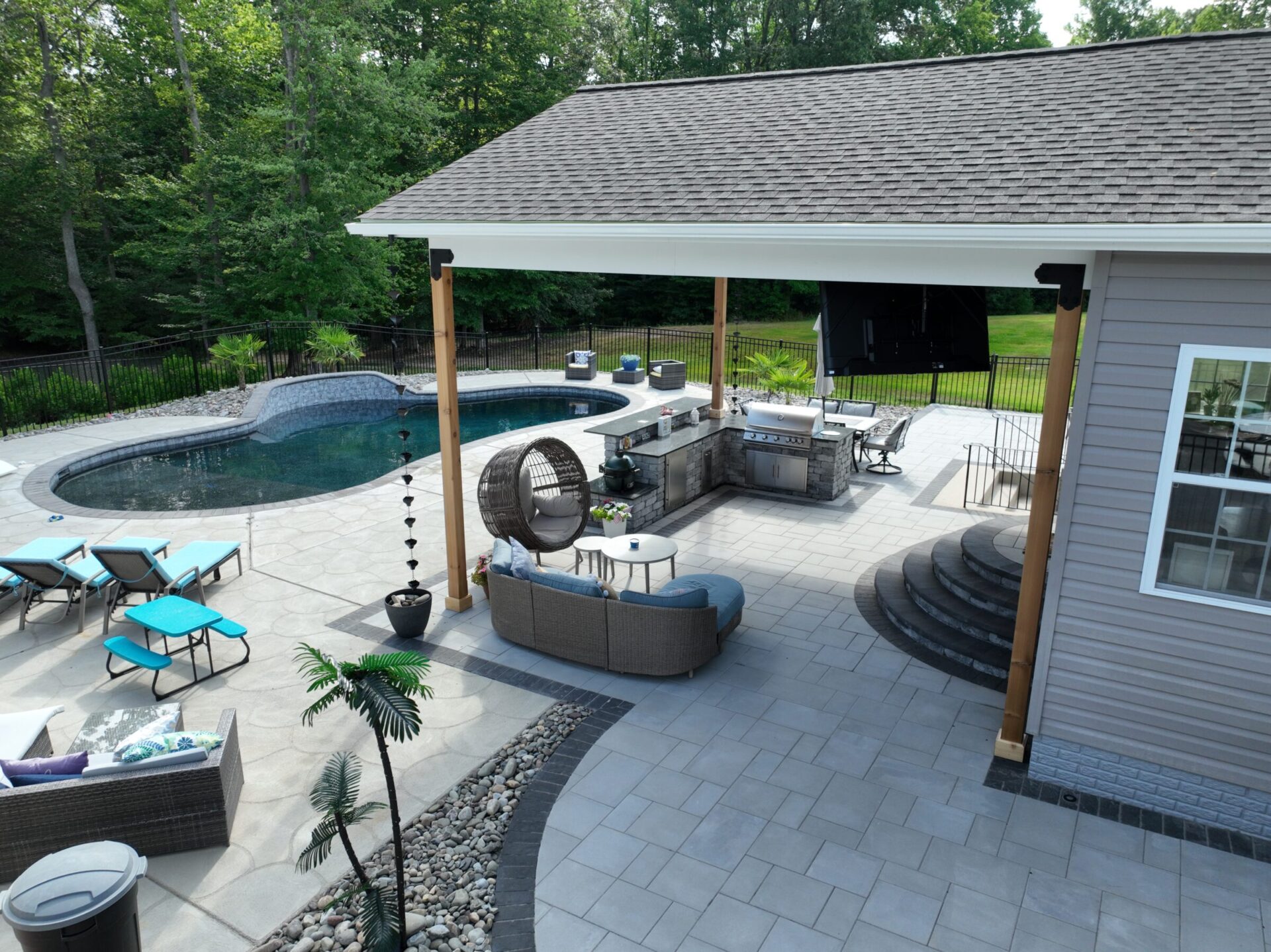 Modern backyard with a swimming pool, patio furniture, and an outdoor kitchen. Surrounded by trees, creating a relaxing and private atmosphere.