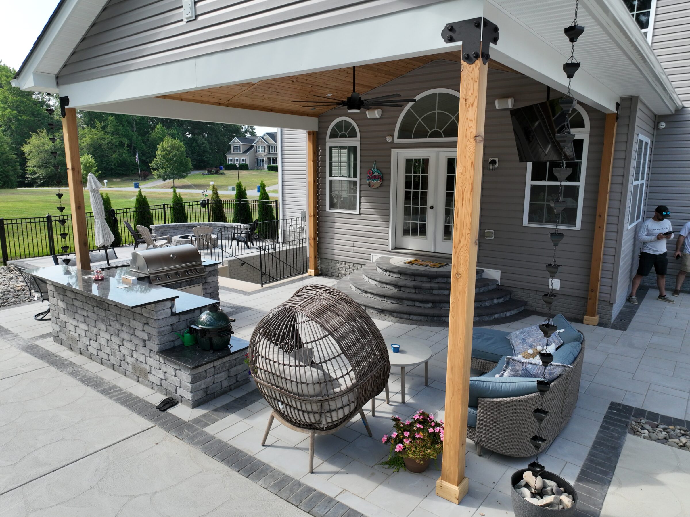 Modern patio with outdoor kitchen, cozy seating, and garden decor. Two-story house in background, person standing nearby, lush green surroundings.