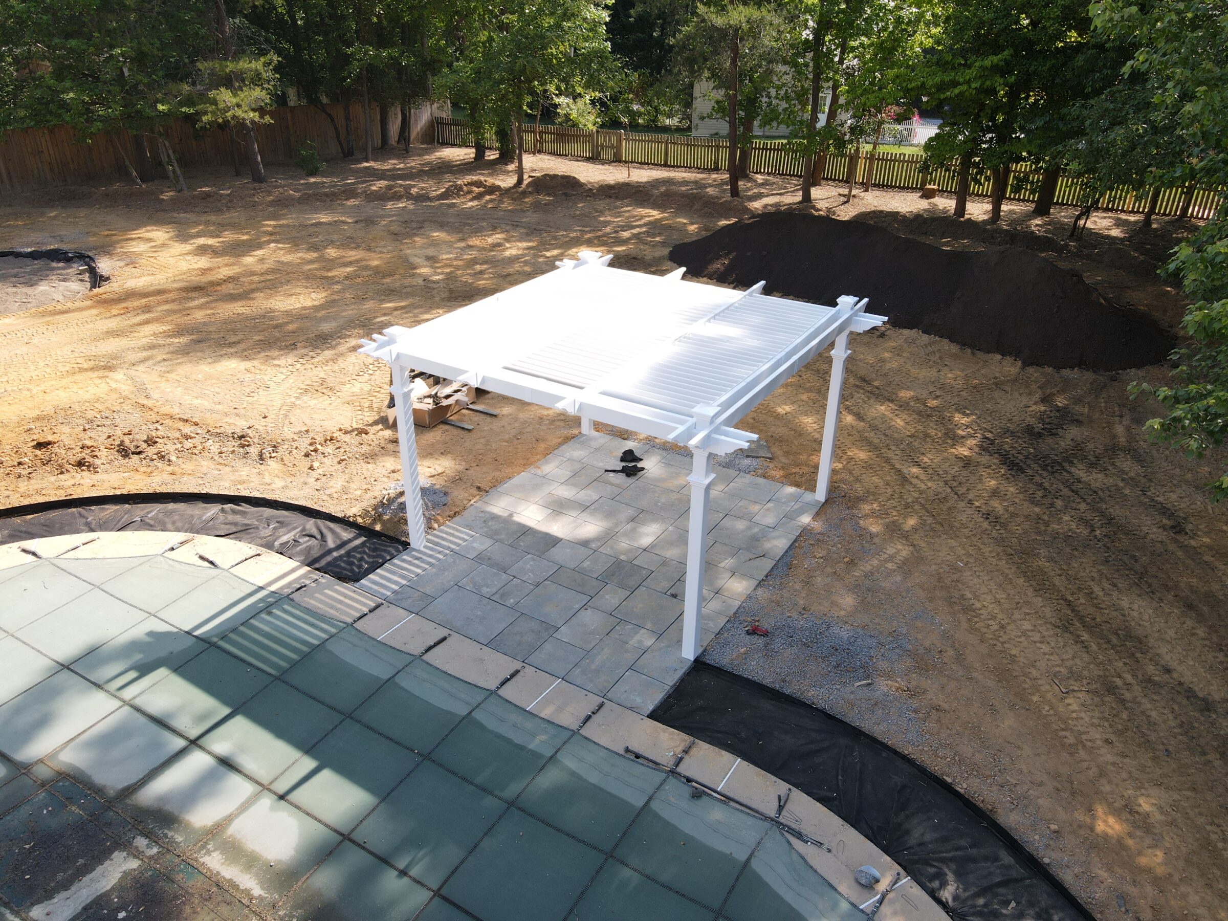 A backyard under construction with a white pergola, paved patio, and groundwork. Trees surround the area with dirt and mulch piles.