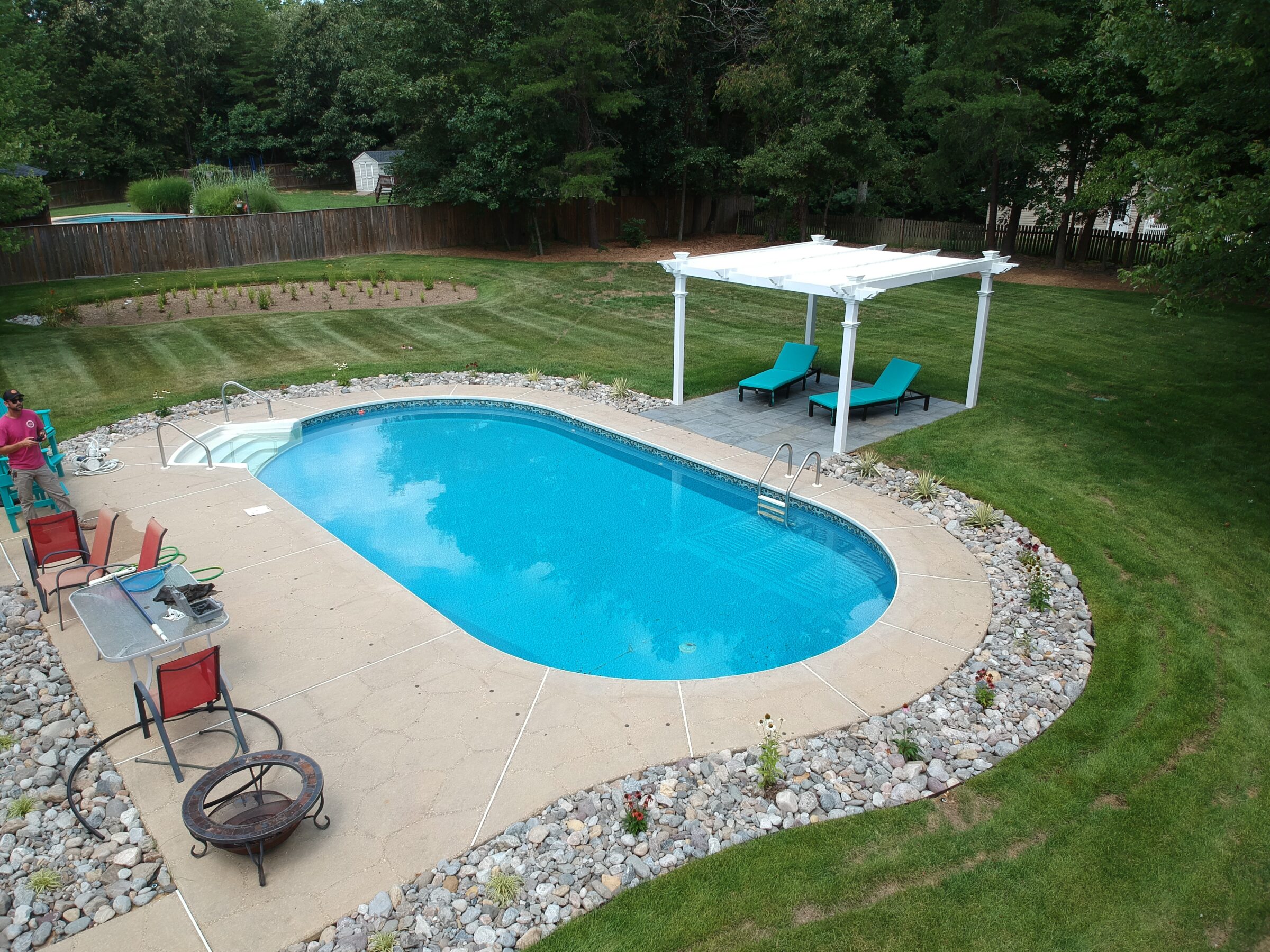 A backyard features a swimming pool, lounge chairs under a pergola, a person near a table, and a grass lawn surrounded by trees.
