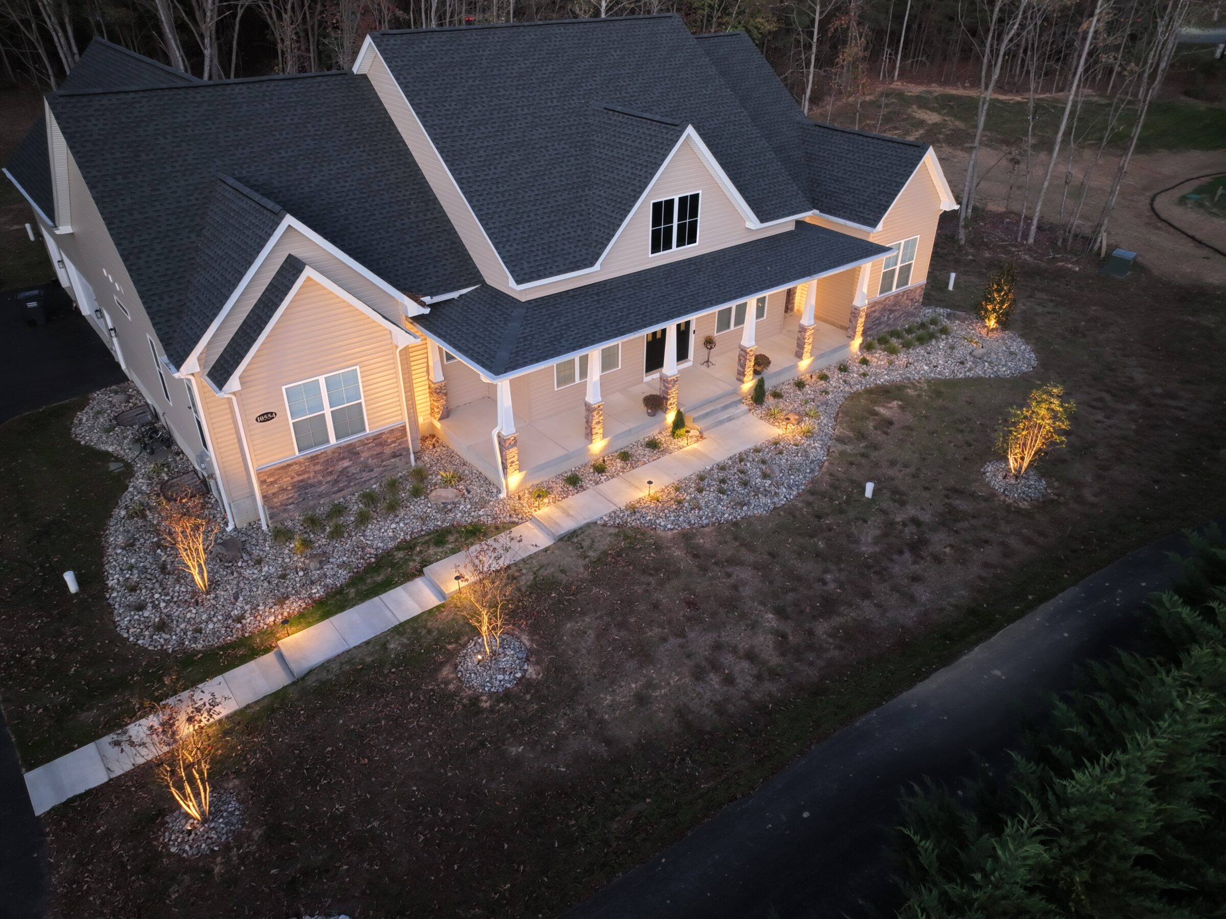 Aerial view of a well-lit modern house with a landscaped garden, set in a wooded area during dusk.
