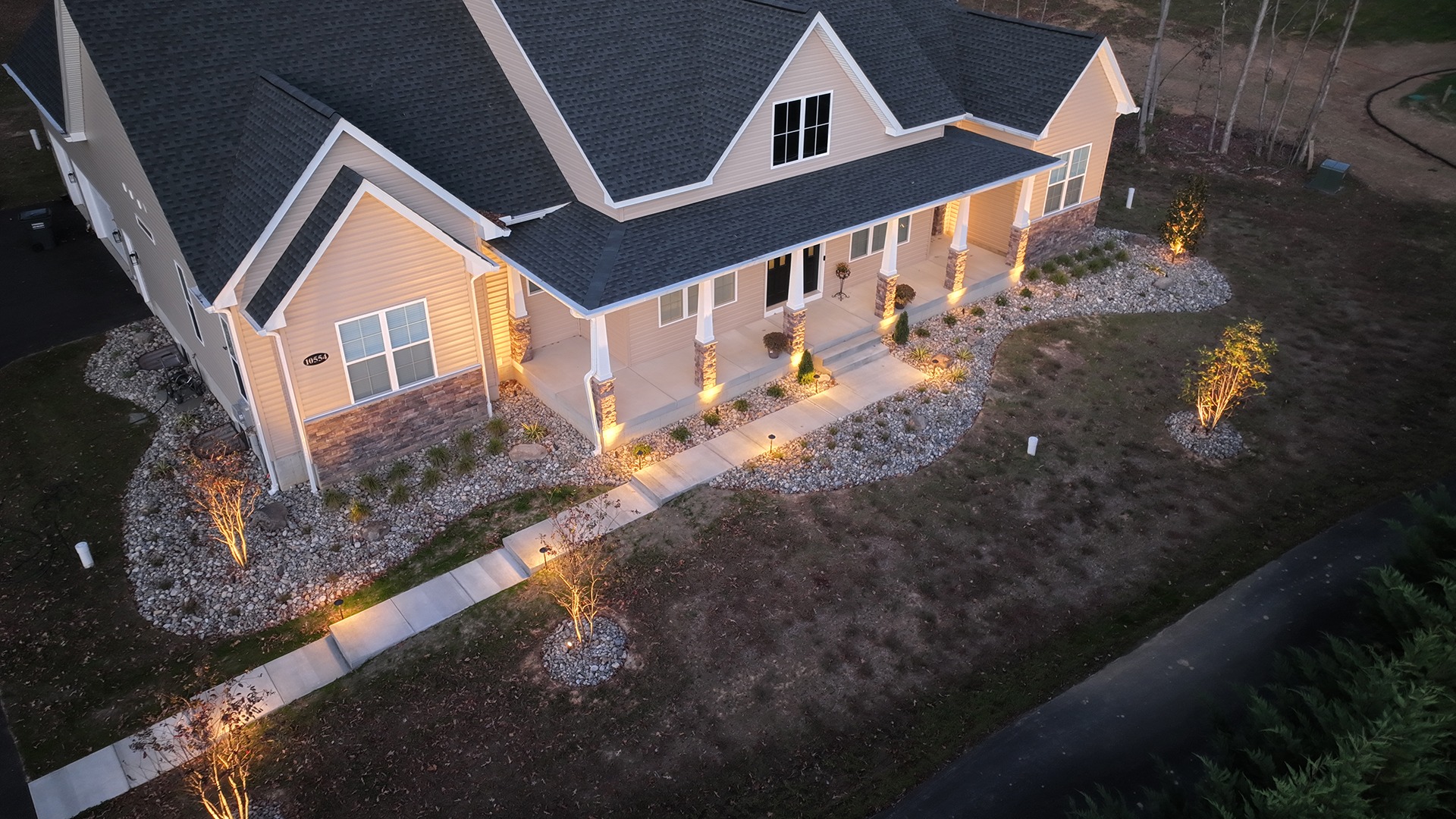 A beautifully illuminated house at dusk, featuring a front porch, landscaped garden with stone paths, and accent lighting highlighting shrubs.