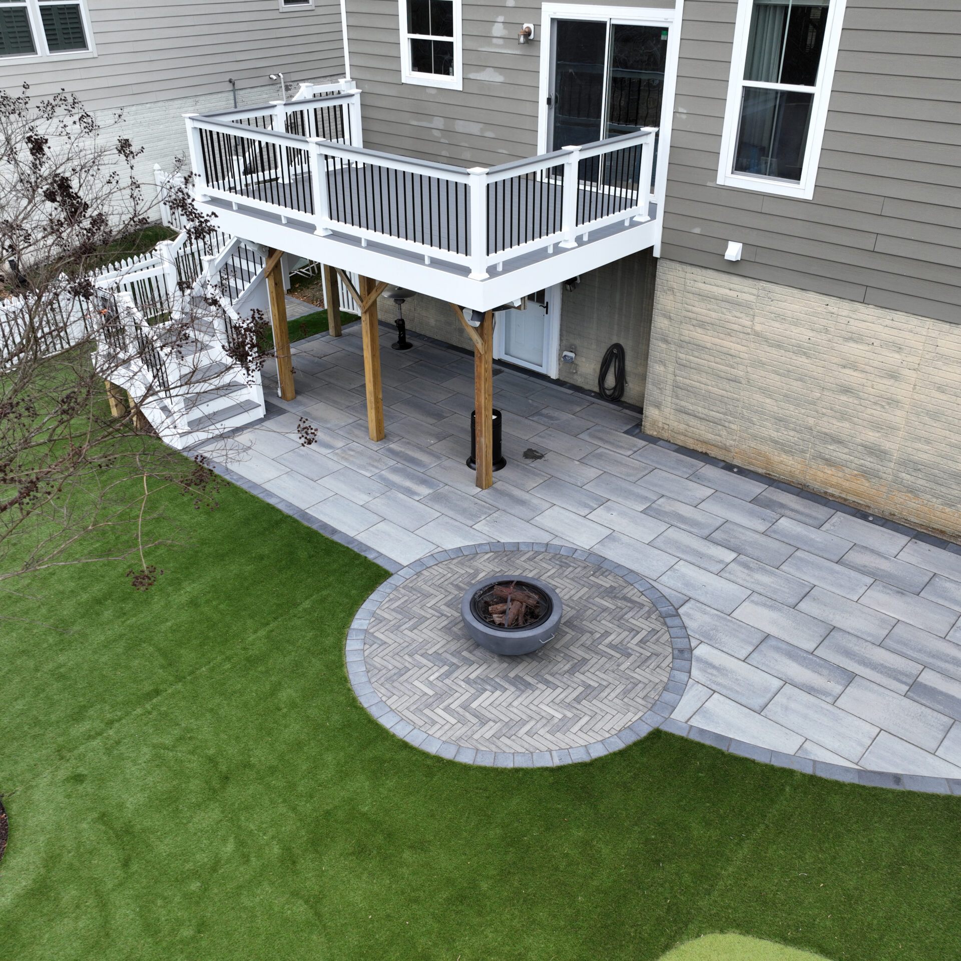 A backyard features a raised deck, paved patio, circular fire pit, and artificial grass, adjacent to a neutral-toned house exterior.