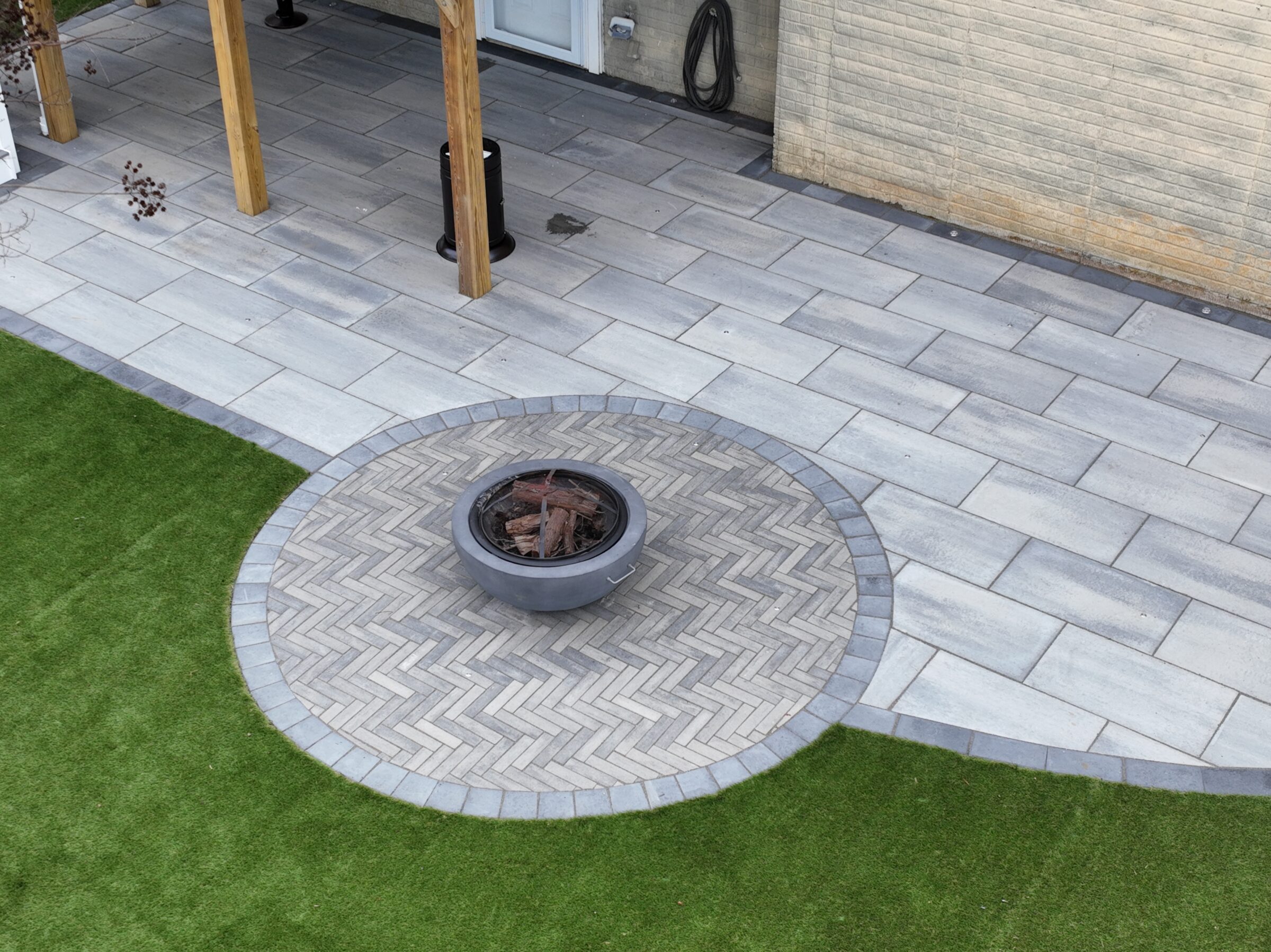 A backyard patio with textured gray tiles, circular fire pit, and artificial green grass, bordered by a brick wall and wooden beams.