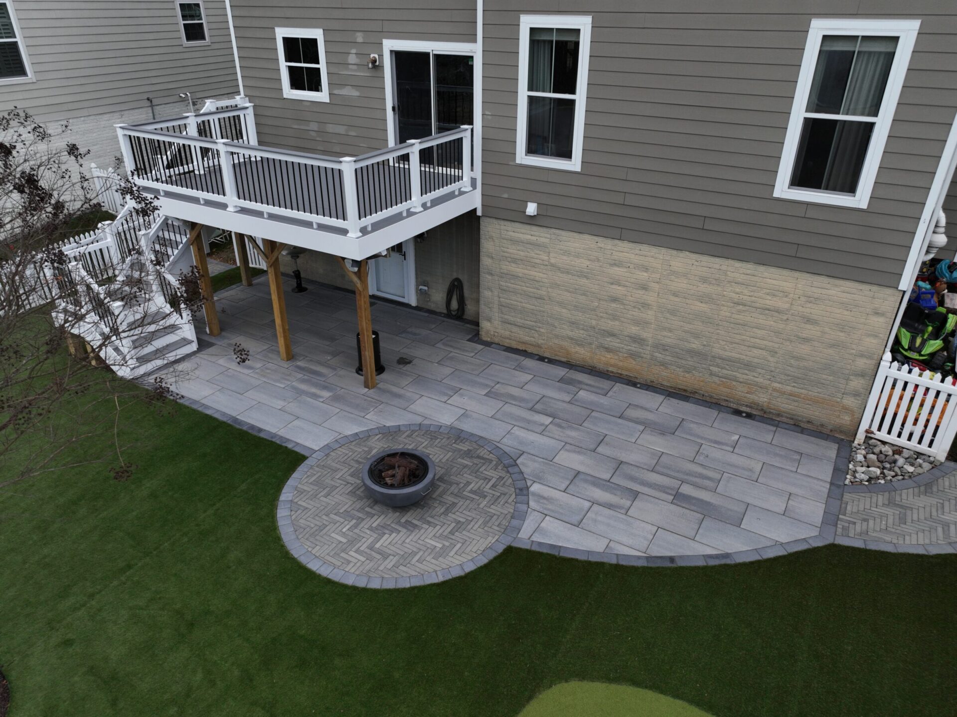 A backyard with a raised deck, patio, fire pit, and artificial grass, adjacent to a house with beige siding and white trim.