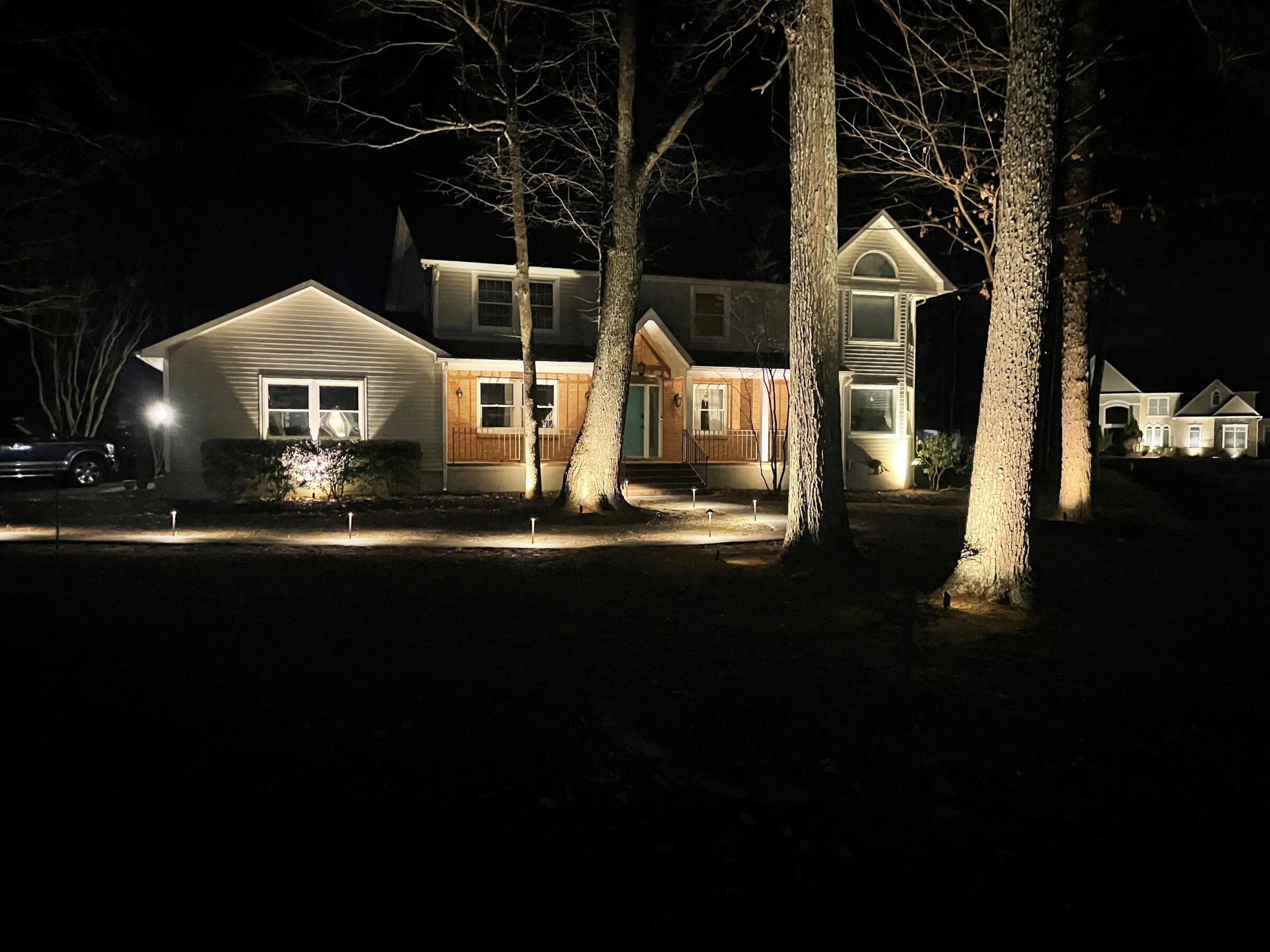 A well-lit house at night, surrounded by tall trees. The scene is tranquil with another illuminated house visible in the background.