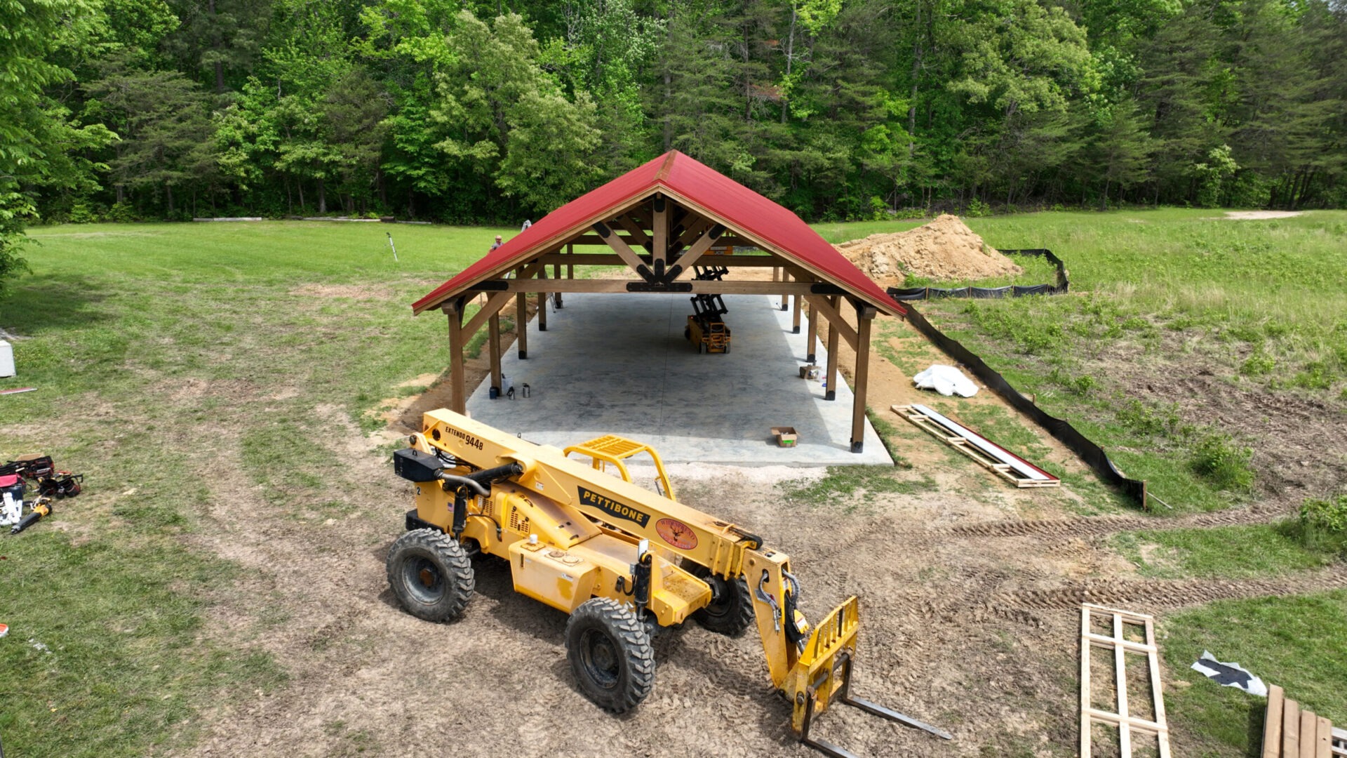 A construction site with a wooden pavilion and red roof, surrounded by trees and grass. Equipment and materials are scattered around.