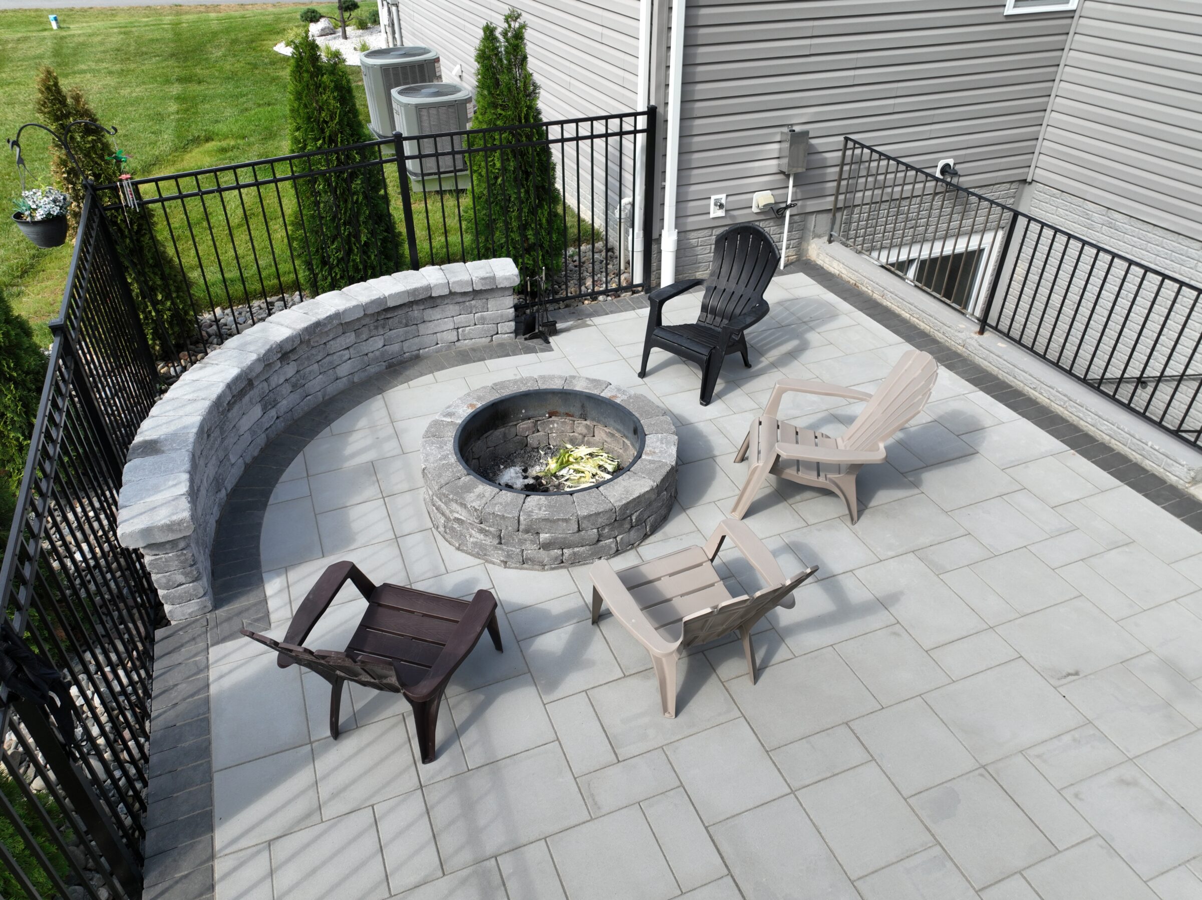 A patio with Adirondack chairs surrounds a stone fire pit, enclosed by metal fencing and a curved stone wall, near a house.