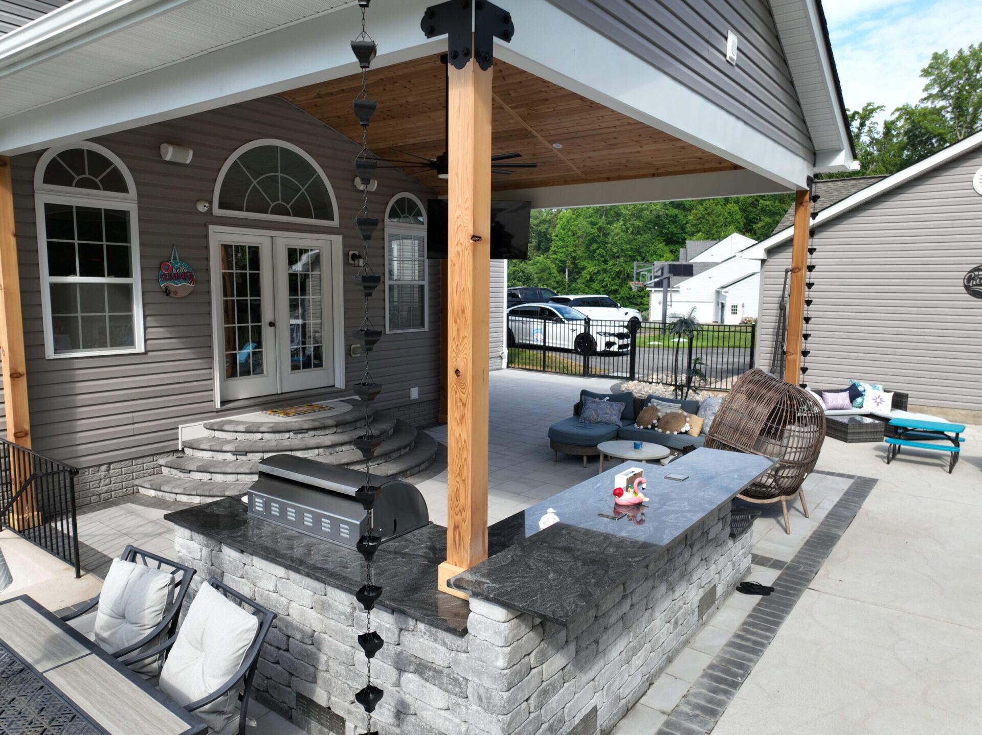 Covered patio with stone seating area, outdoor grill, modern furniture, and decorative elements. Suburban background with a parked vehicle and houses.