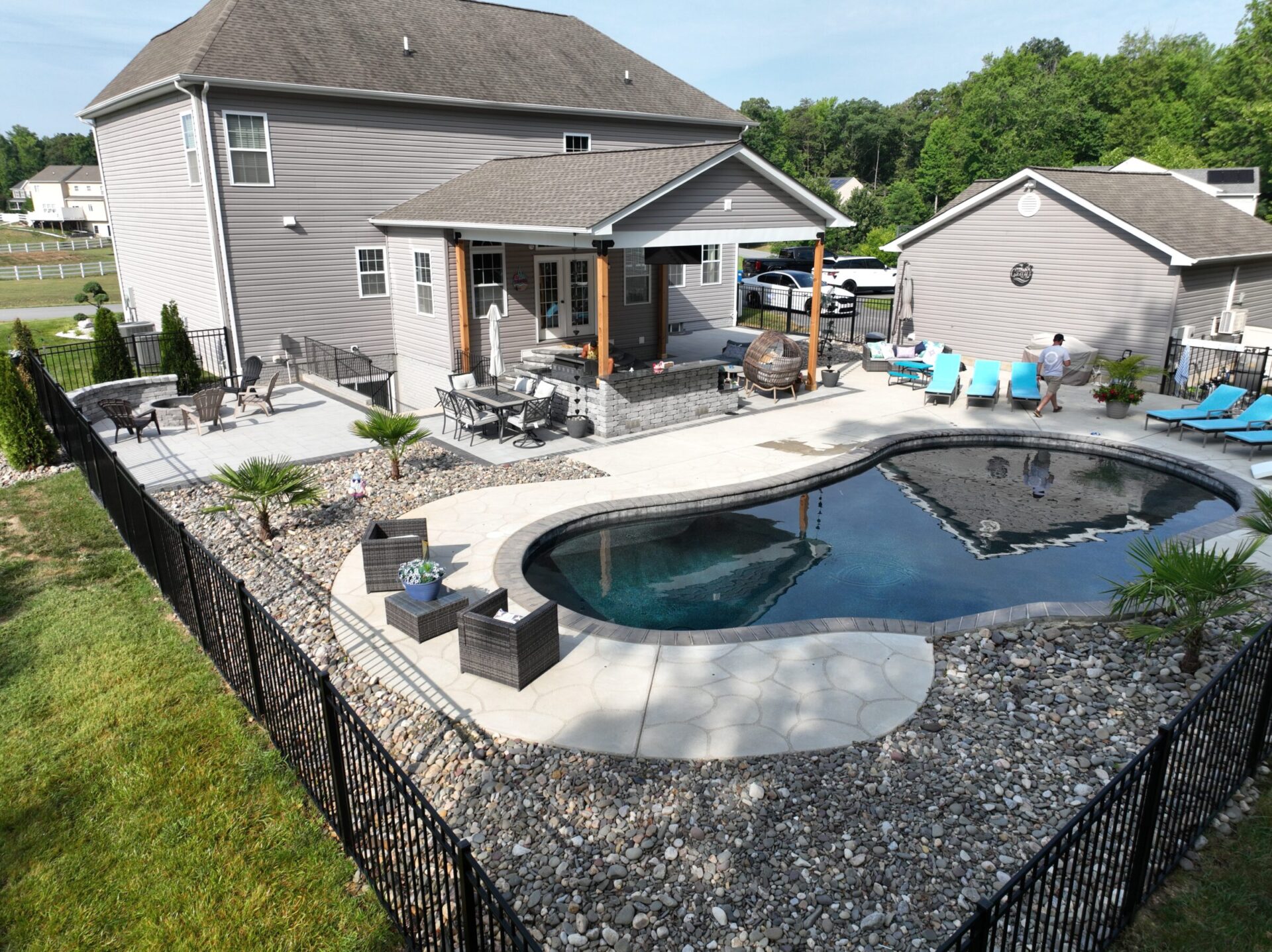 Modern backyard features a pool, patio seating, and a person near lounge chairs. House and greenery are visible in the background.