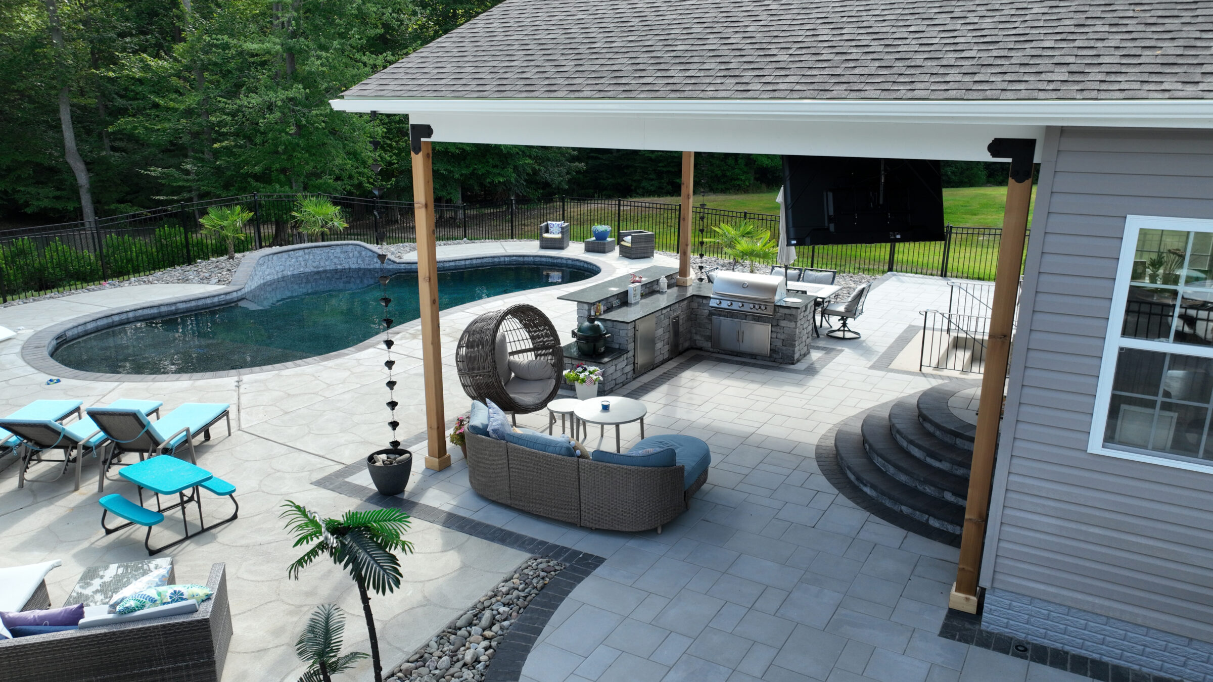 Modern backyard with a pool, outdoor kitchen, and seating area, surrounded by trees. Features lounge chairs and a partially covered patio.