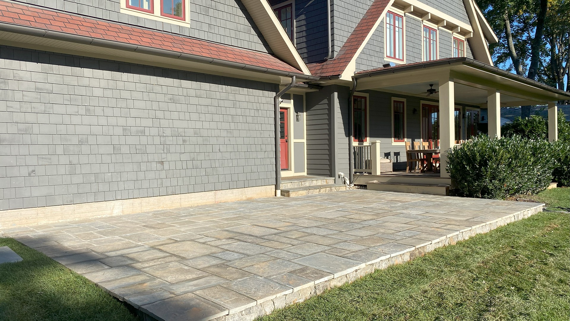 A house with gray shingle siding features a stone patio, covered porch, and outdoor seating surrounded by greenery on a sunny day.