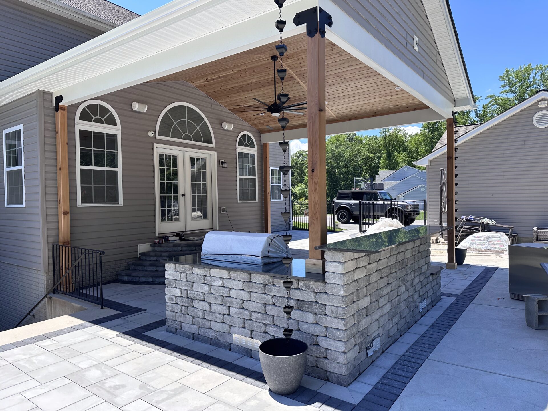 A residential patio with stone bar, covered seating area, and modern design. Backyard features a parked vehicle and trees in the background.