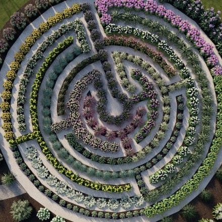 Circular garden maze with vibrant, colorful flowers arranged in geometric patterns, surrounded by greenery. Pathways create an intricate, beautiful design. No people visible.