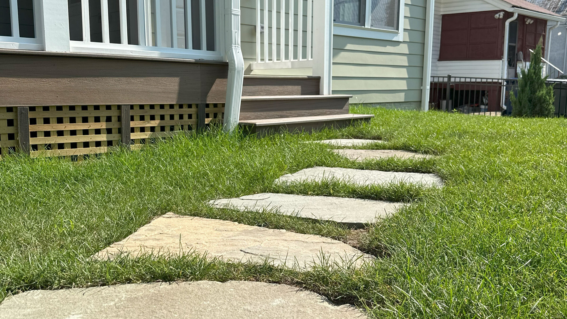 Stone pathway leads to a porch amidst green grass and neighboring houses. Steps and railing enhance the home entrance’s inviting feel.