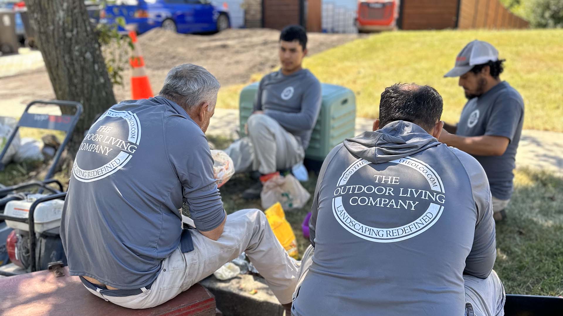 Four people in matching company attire sit outdoors, taking a break with equipment nearby on a sunny day, under a tree.