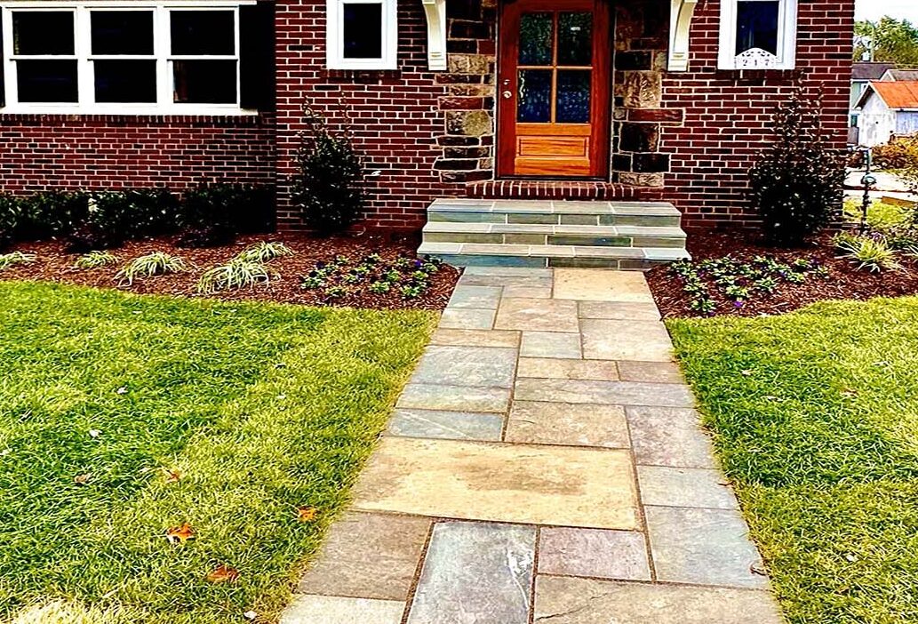 A charming brick house with dark shutters, manicured lawn, and stone pathway, surrounded by greenery and neighboring homes under a clear sky.