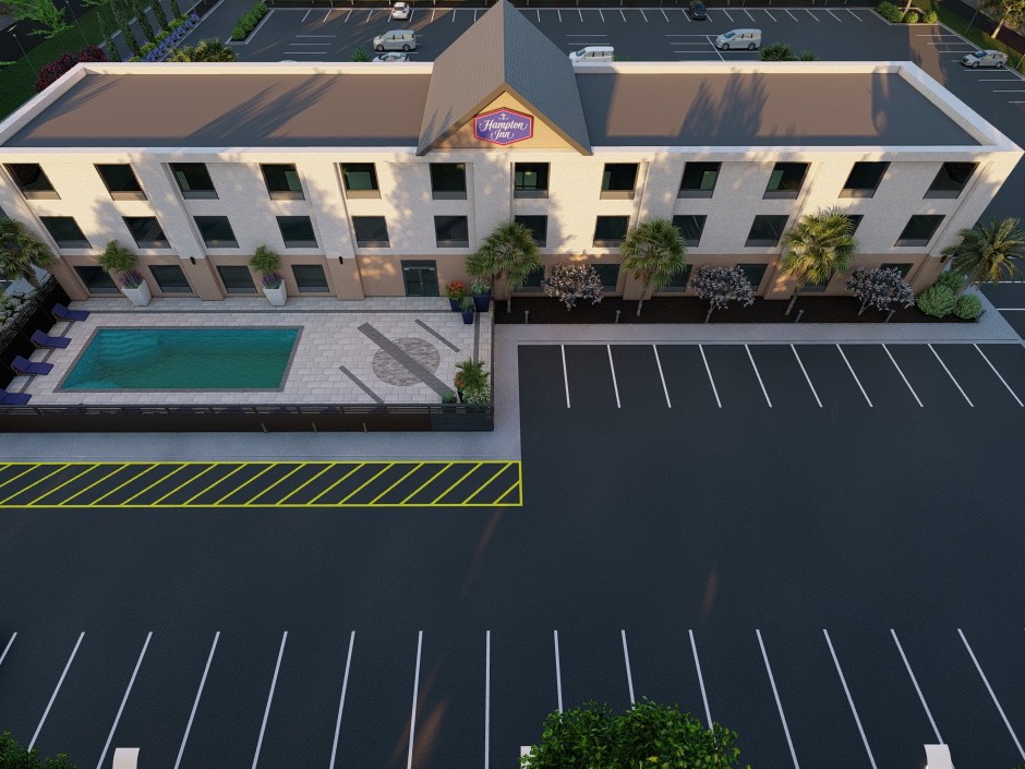 Three-story Hampton Inn with parking lot, swimming pool, palm trees, and outdoor seating area. Minimal vehicles visible, clear sky reflects evening shadows.