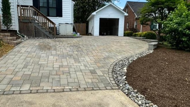 A neatly paved driveway leads to a white garage beside a white house, with a landscaped garden featuring mulch and decorative stones.