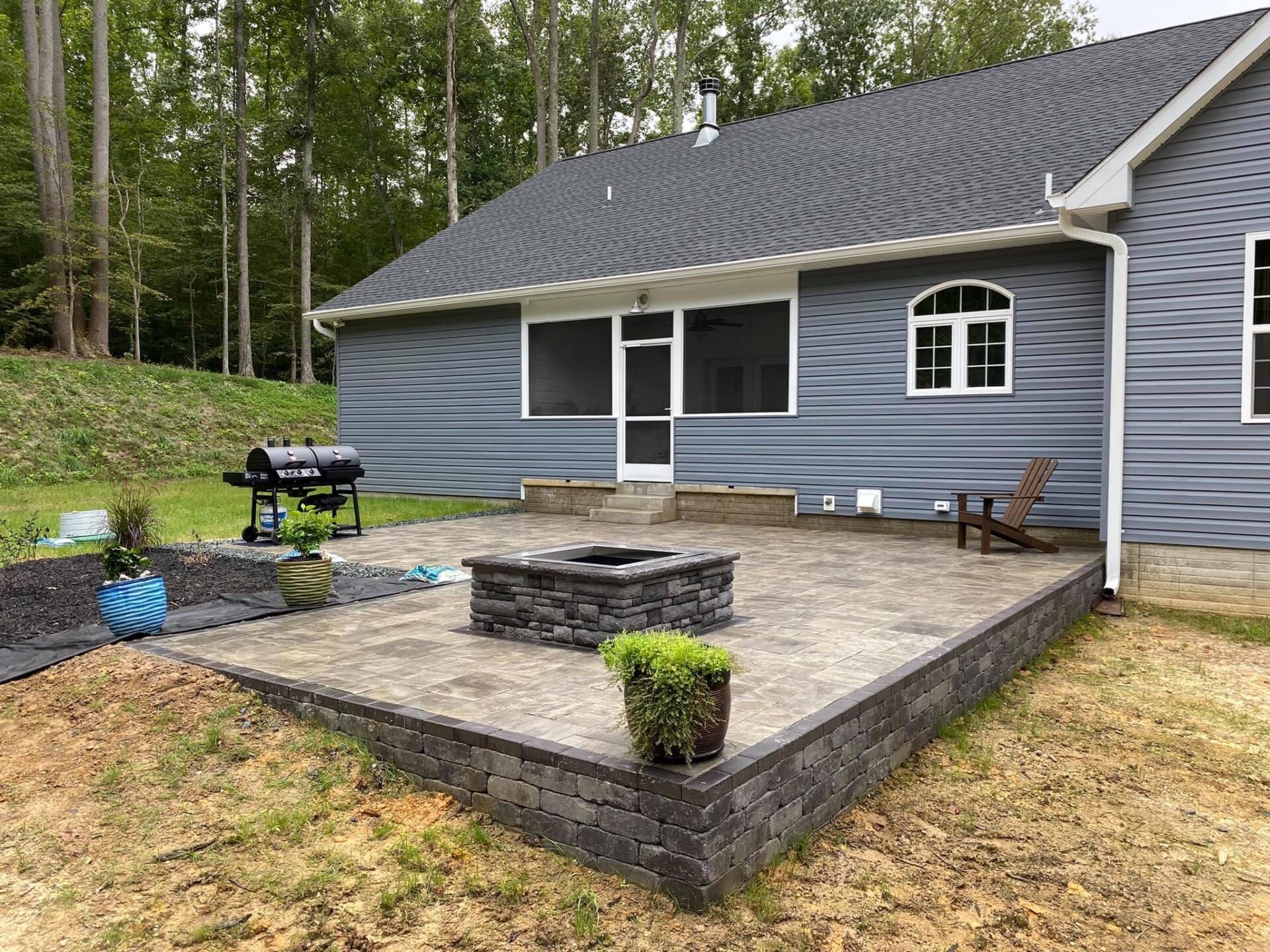 A modern house with a stone patio, grill, and fire pit, surrounded by trees. Includes two potted plants and an empty chair.