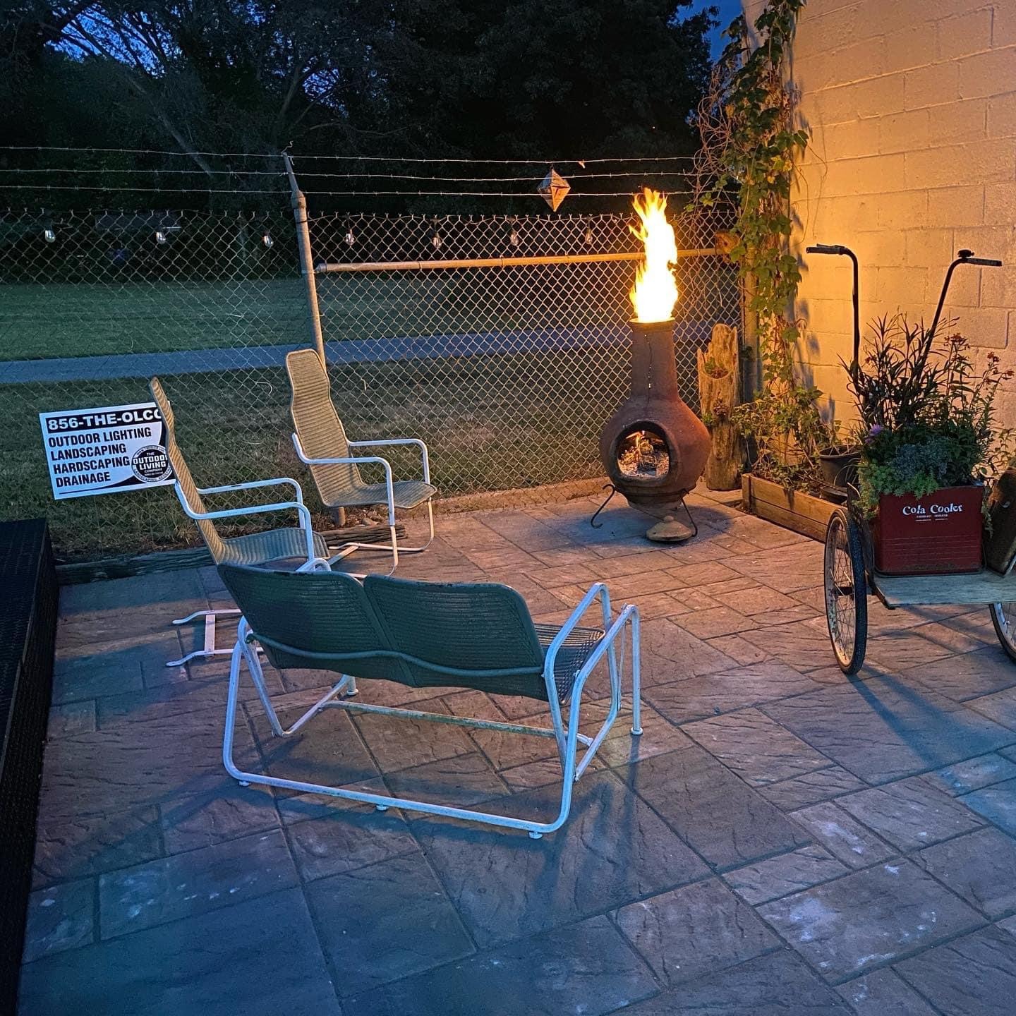 A patio with chairs and a fire pit beside a bicycle planter, enclosed by a chain-link fence at dusk.