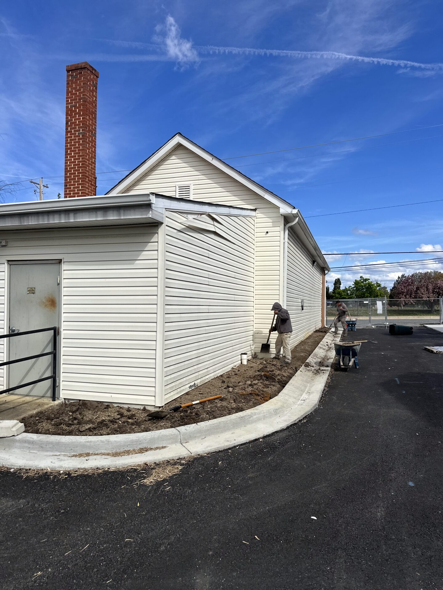 A small, simple building with two people working outside, surrounded by clear blue skies and an asphalt driveway.