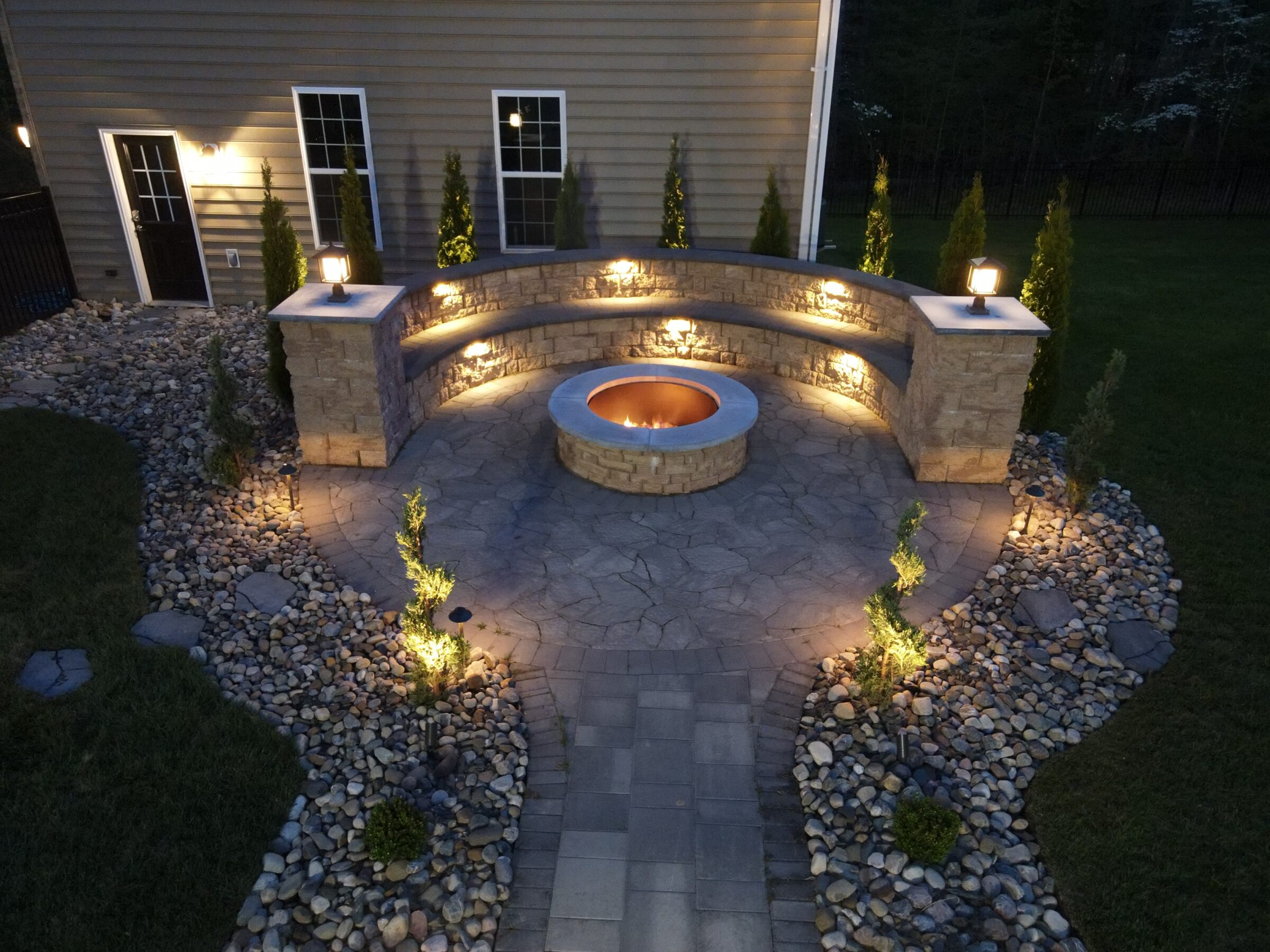 Outdoor stone patio with circular fire pit, surrounded by illuminated landscaping, pebbled pathways, and adjacent building wall featuring windows and a door.