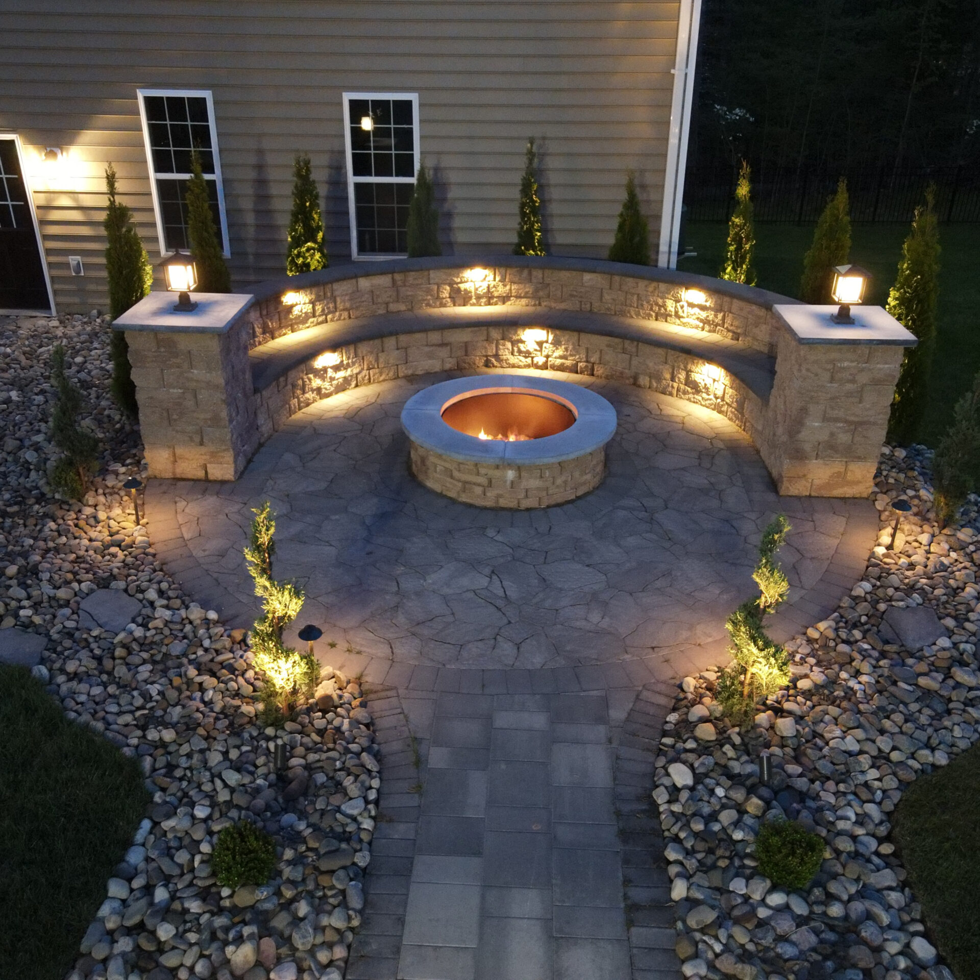 A cozy outdoor firepit area with circular seating, surrounded by illuminated stonework and pebbles, adjacent to a house in evening light.