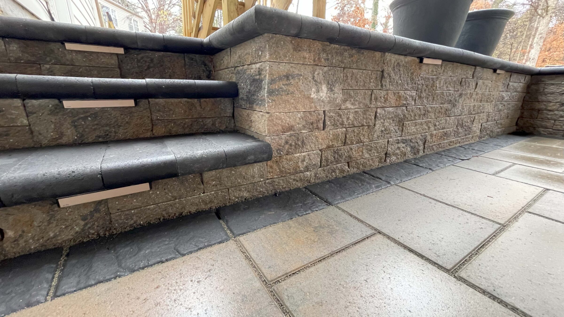 Stone steps and retainment wall with modern, black edging. Paved patio area with large planters, surrounded by trees in the background.