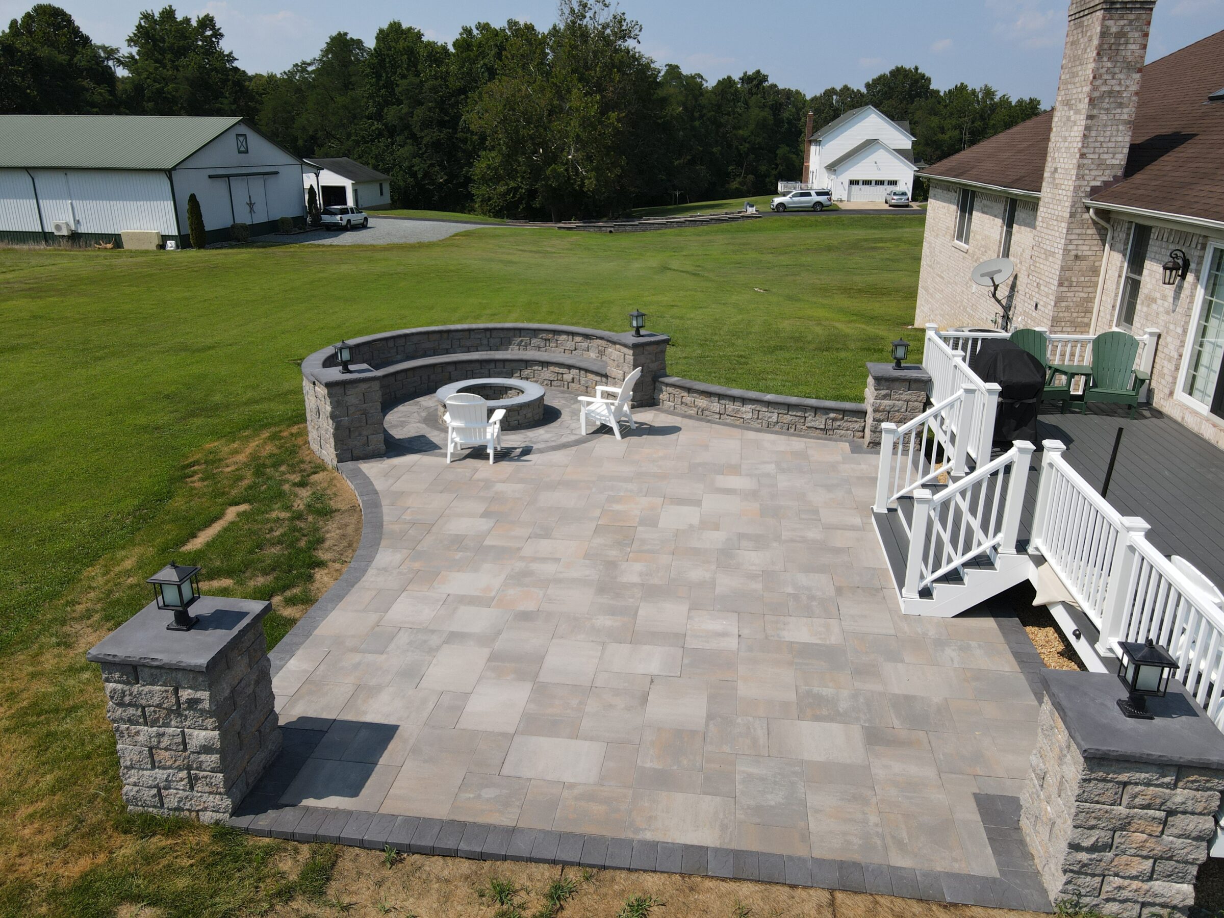 A spacious backyard with a stone patio, seating area, and steps. Green fields and outbuildings surround a residential house.