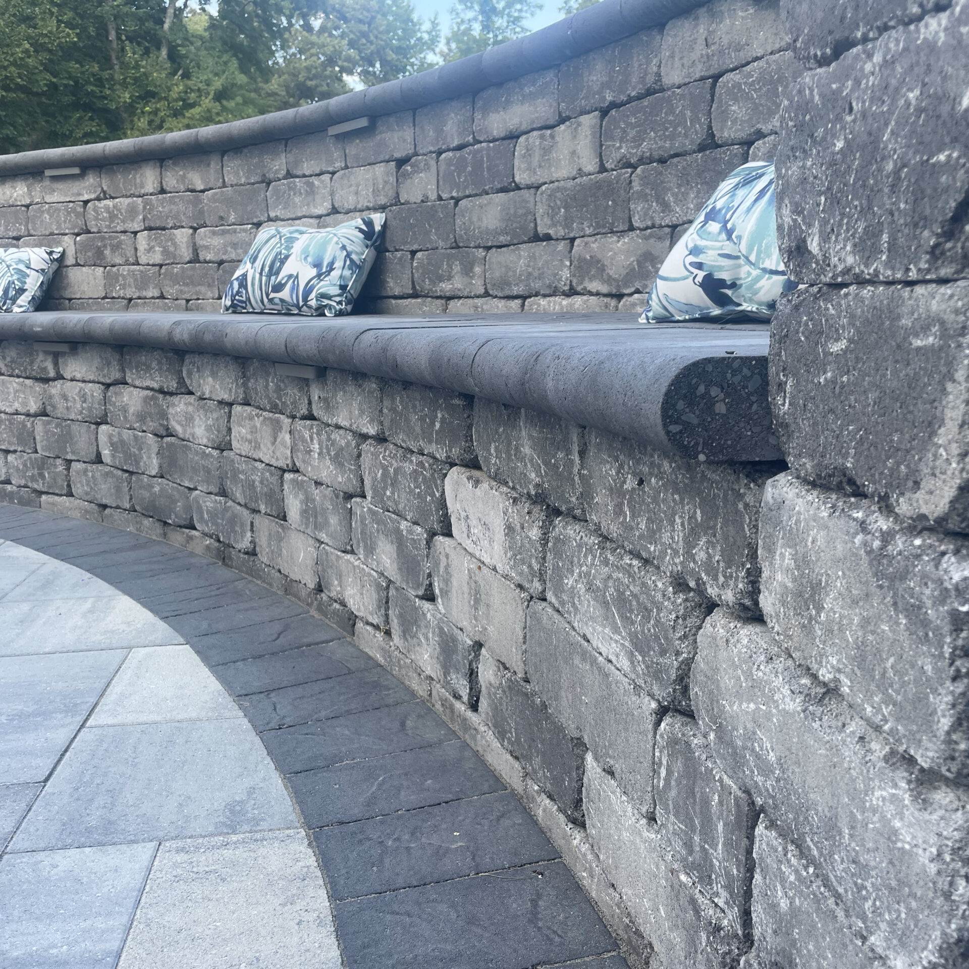 Curved stone bench with patterned cushions, set outdoors. Surrounded by trees, the seating has a rustic appearance and offers serene relaxation.