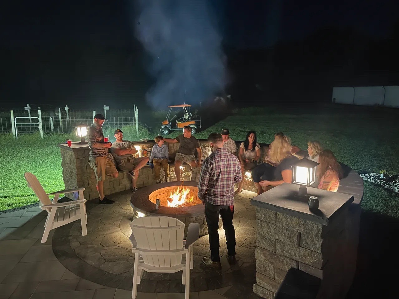 A group of people gather around a fire pit at night, seated and standing, with a backdrop of fenced grass.